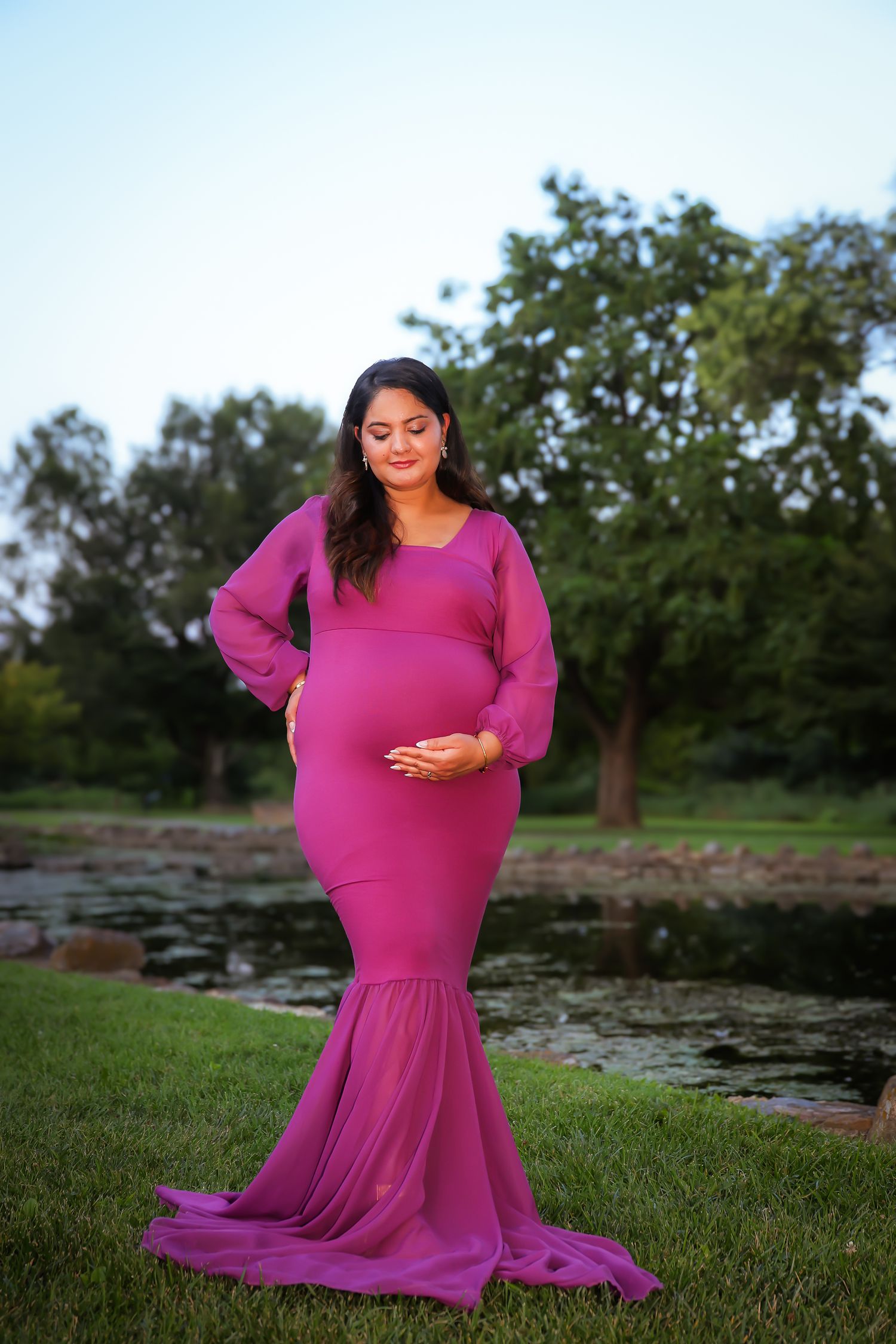 A person in a flowing magenta dress poses gracefully on grassy terrain near a pond during an outdoor photo session.