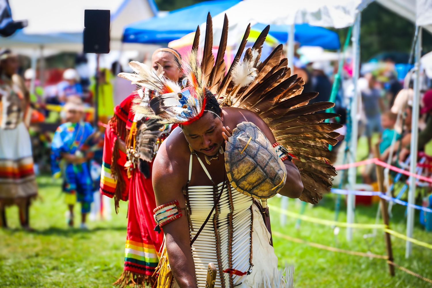 Native American dancers in traditional regalia perform at an outdoor cultural festival with colorful feathered headdresses.