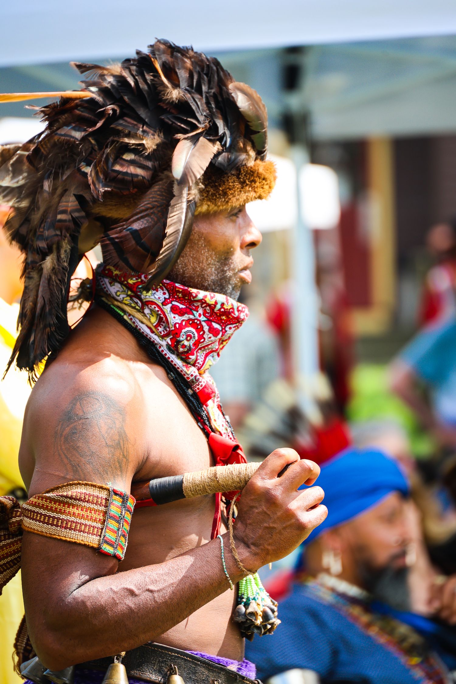 Indigenous performer wearing traditional feather headdress and red bandana plays wooden flute at cultural festival.