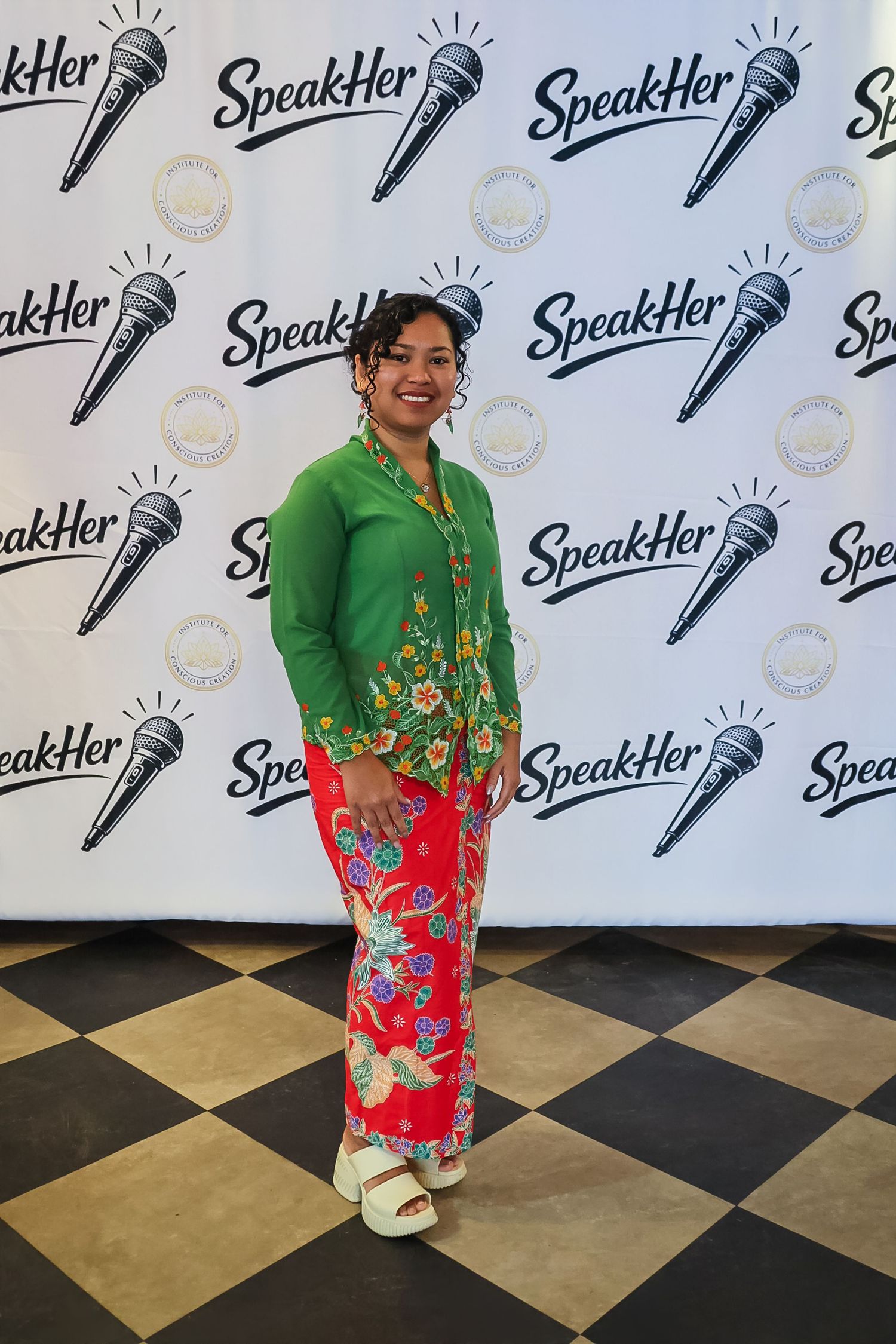Woman in vibrant green floral jacket and red pants poses at SpeakHer event backdrop.