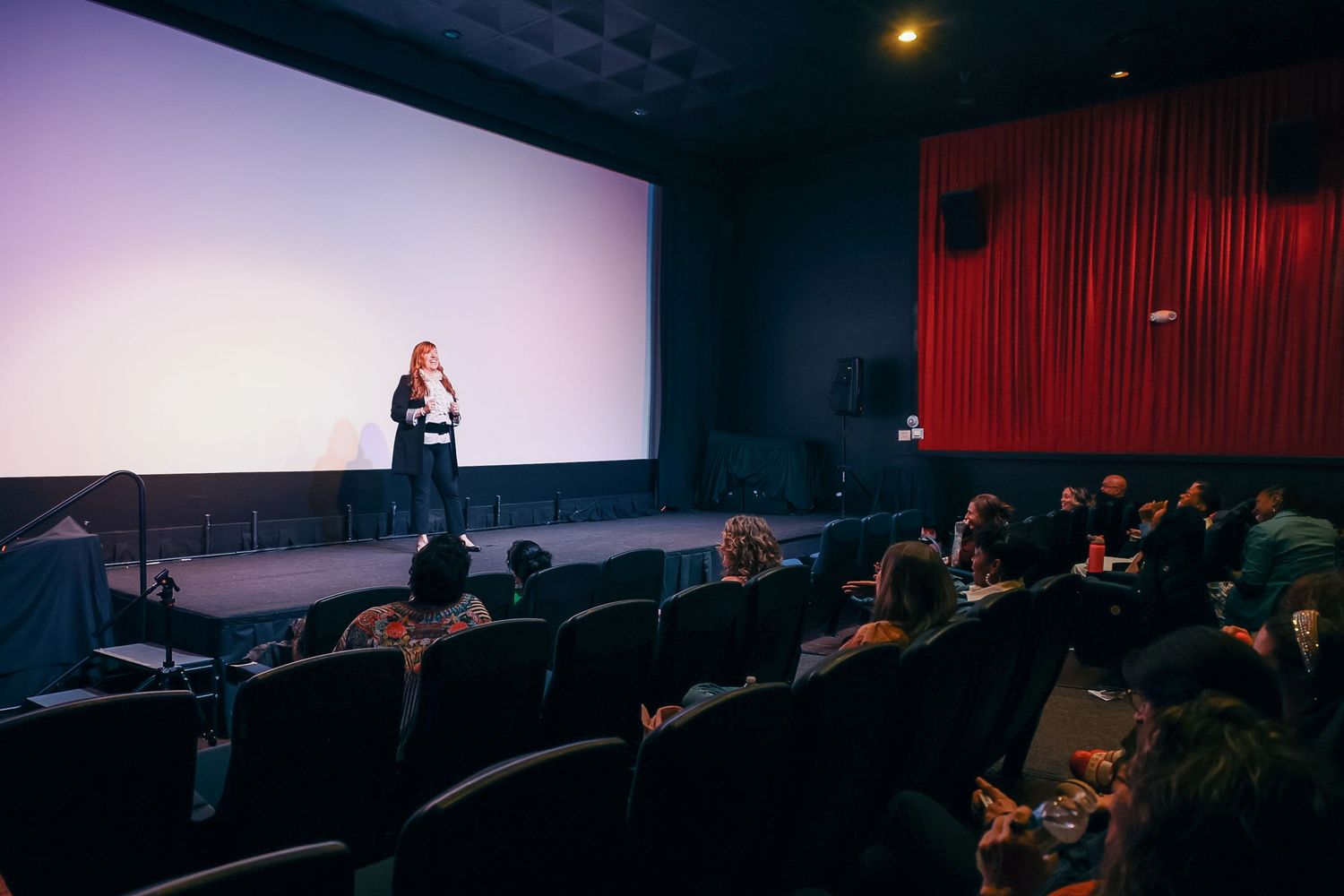 Woman presenting on stage in a cinema with a large white screen, red curtains, and an audience seated in dark theater chairs.
