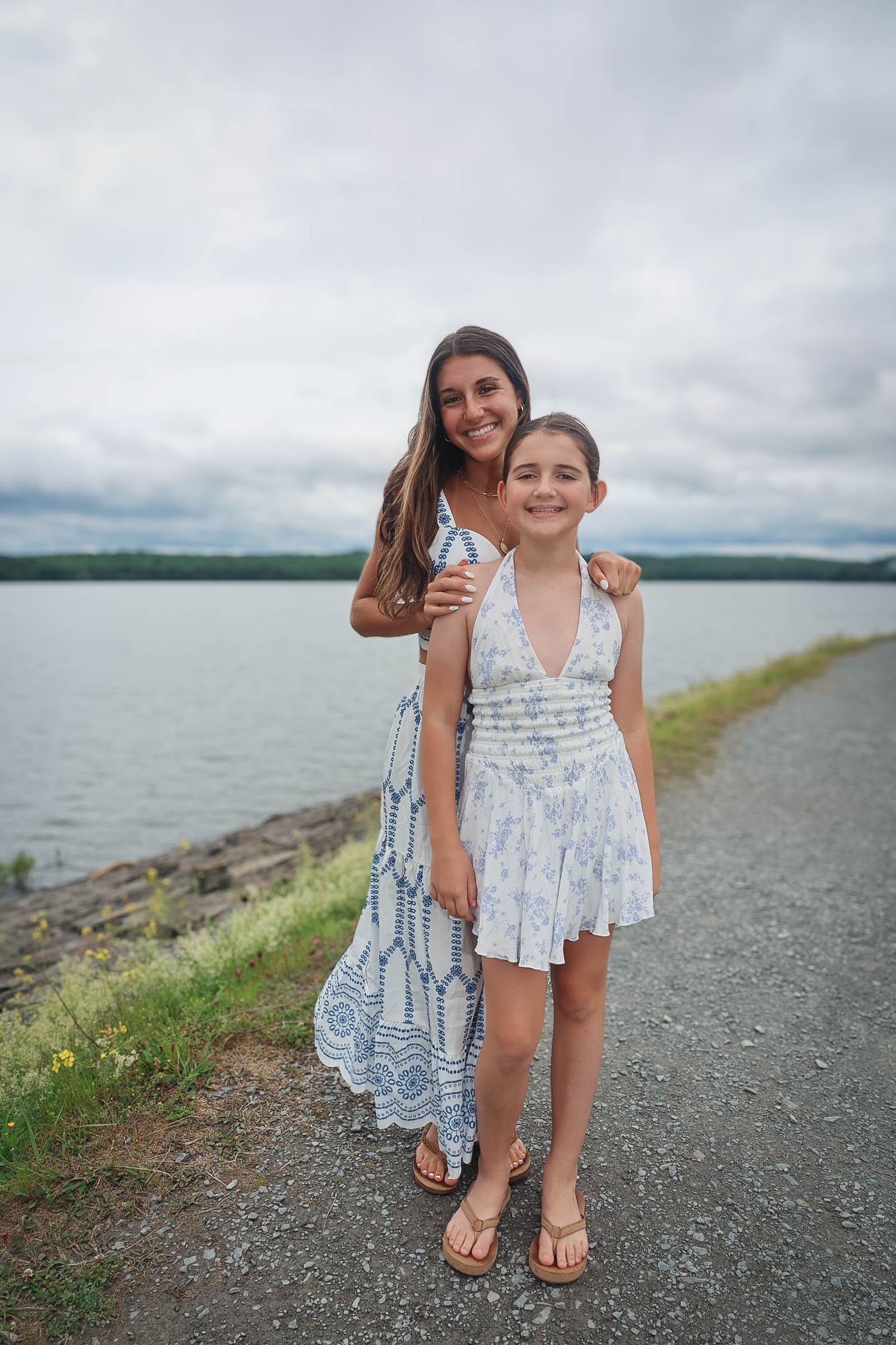 Two girls in floral sundresses pose together on a lakeside path under a cloudy sky.