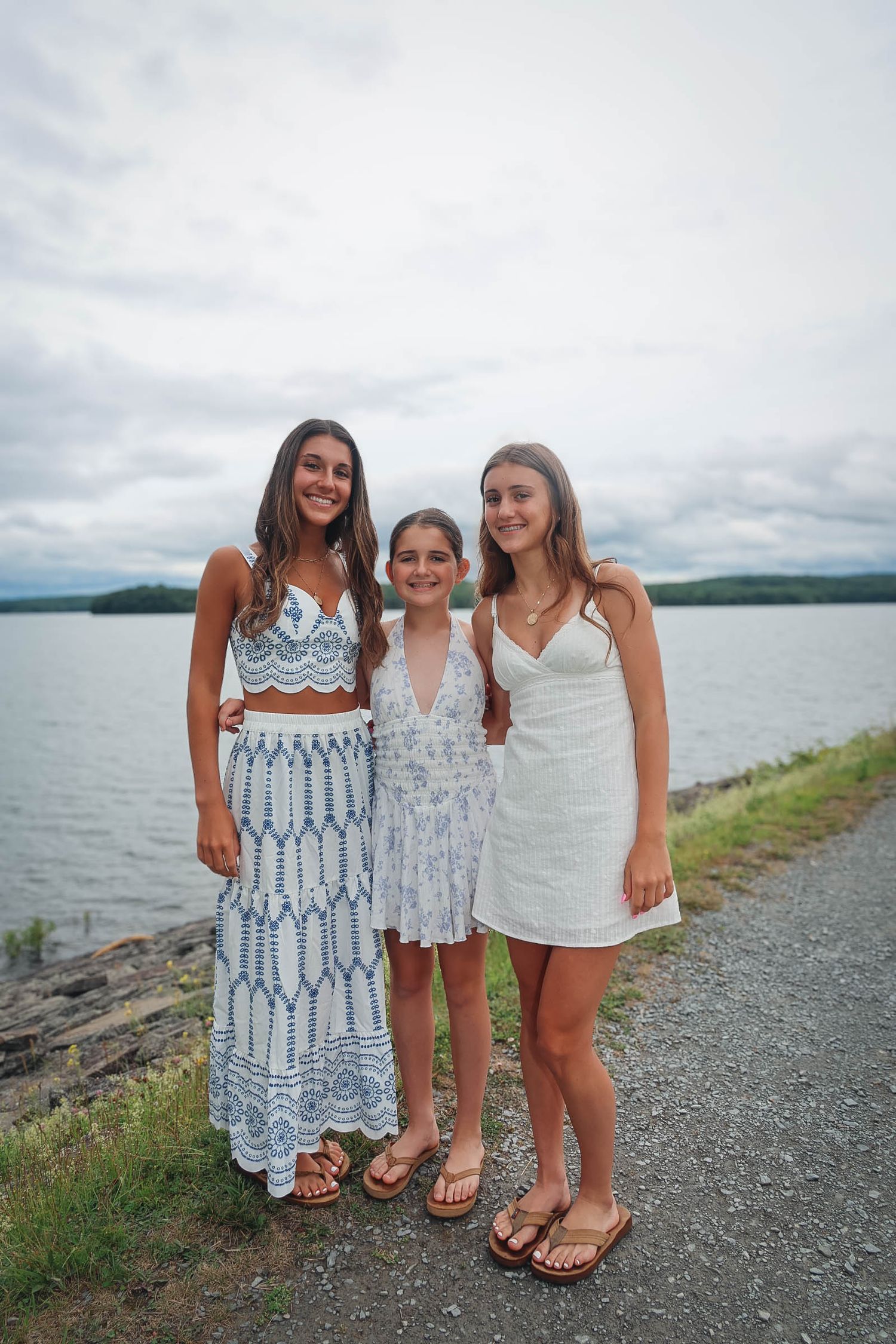 Three teenage girls in white summer dresses pose together smiling by a lakeside on a cloudy day.