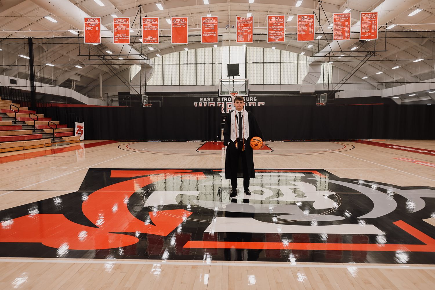 Person standing at center court of a basketball gymnasium with red and black team logo on hardwood floor.