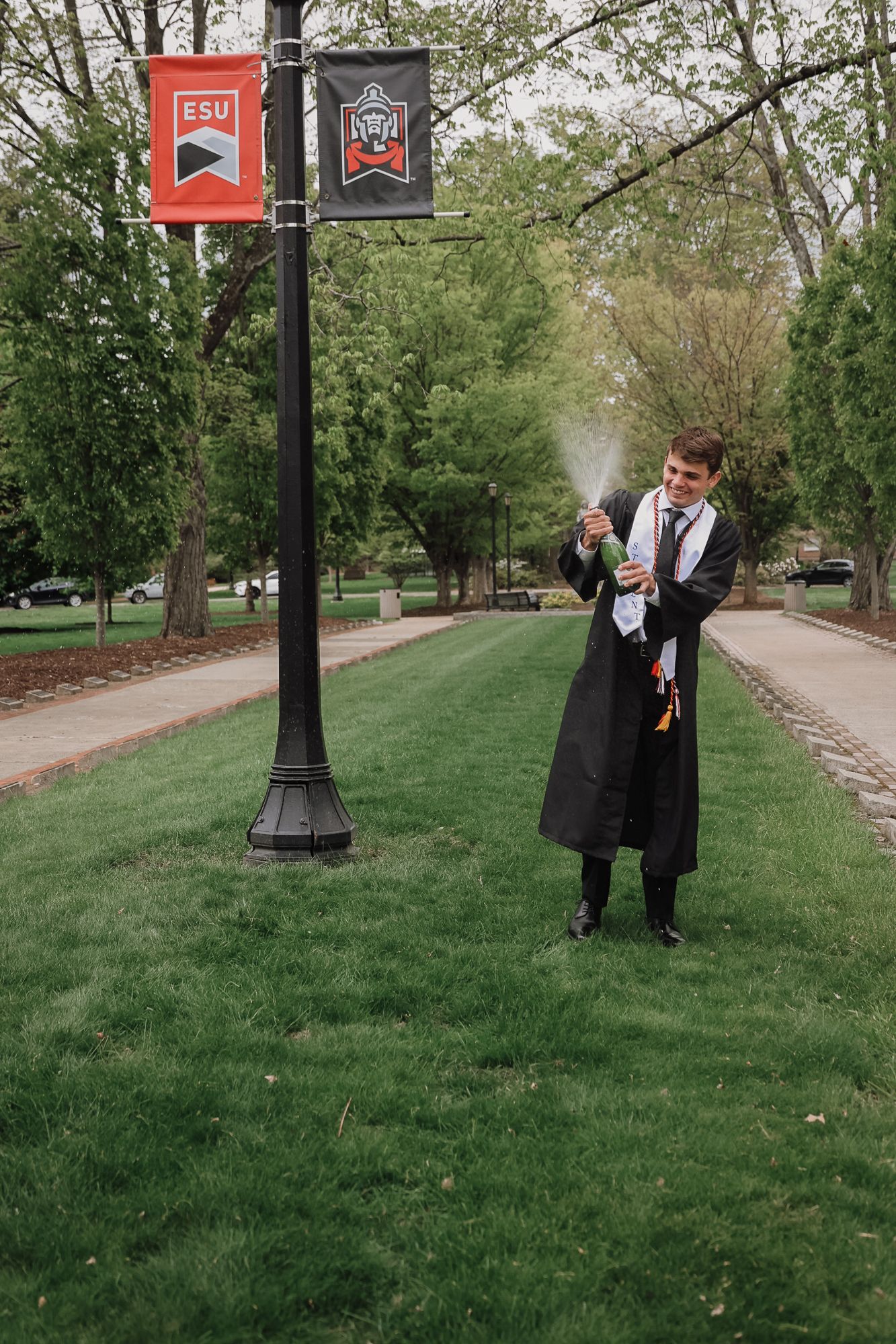 Graduate in black gown pops champagne bottle on ESU campus lawn during outdoor graduation celebration.