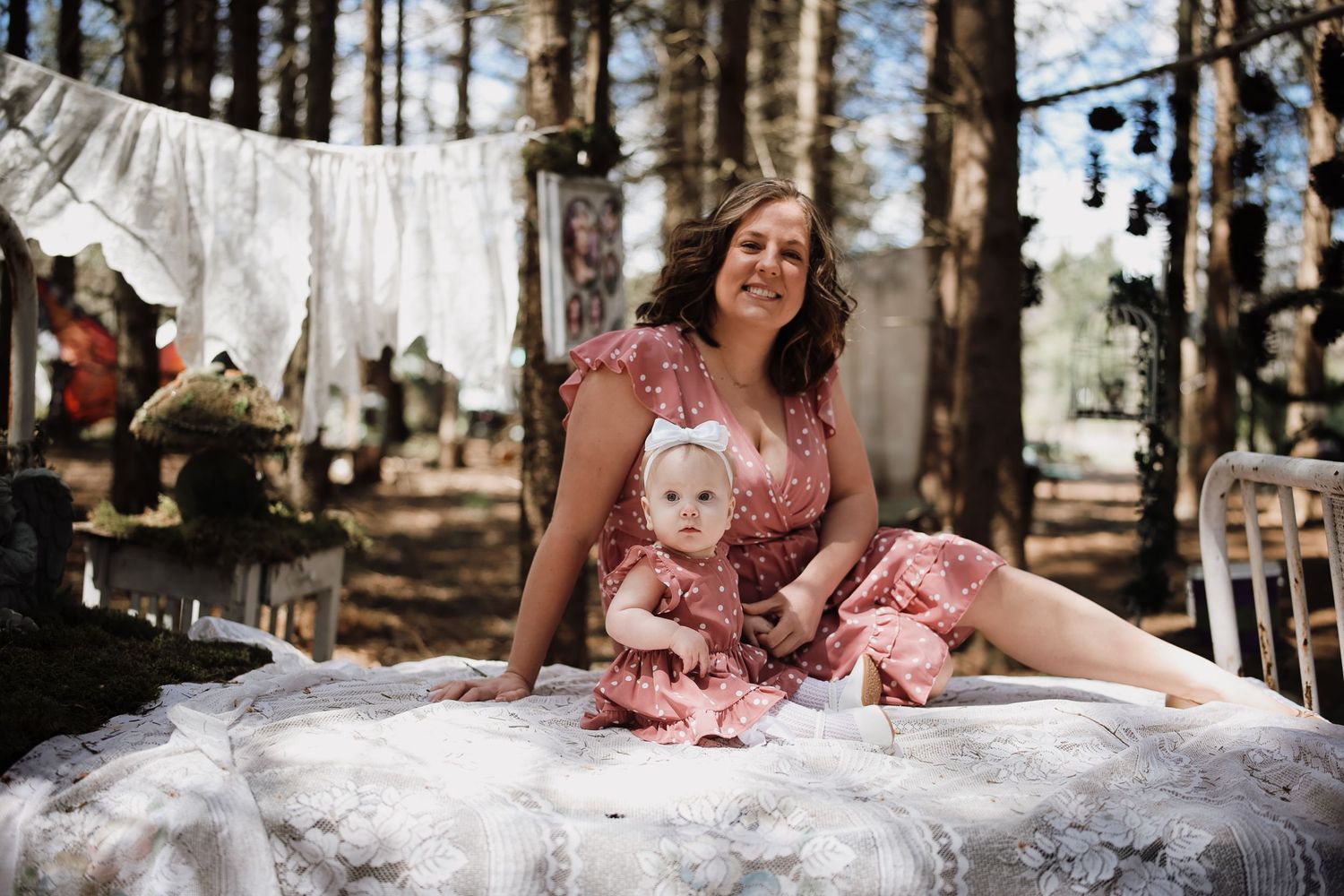 Woman in pink dress poses with baby girl in matching outfit on white lace blanket in rustic woodland setting.