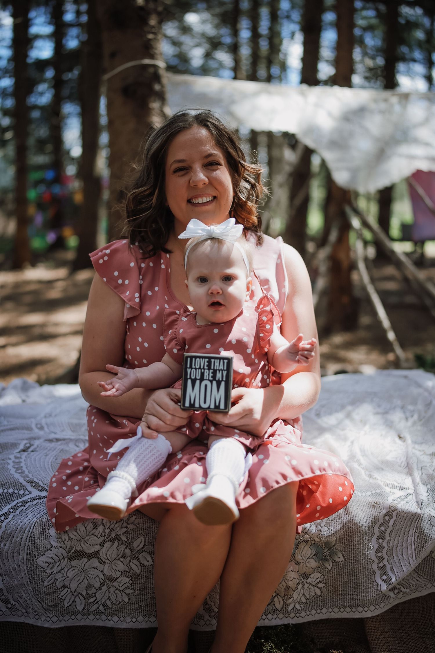 A smiling mother in a pink dress holds her baby girl, who clutches an 'I Love That You're My Mom' book.
