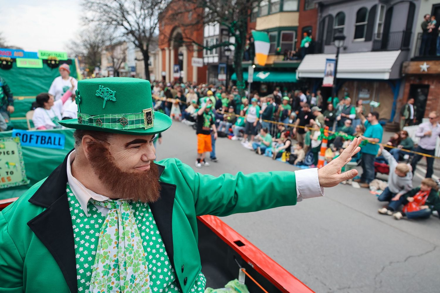 Man dressed as a leprechaun in green suit waves to crowd at a St. Patrick's Day parade on a city street.