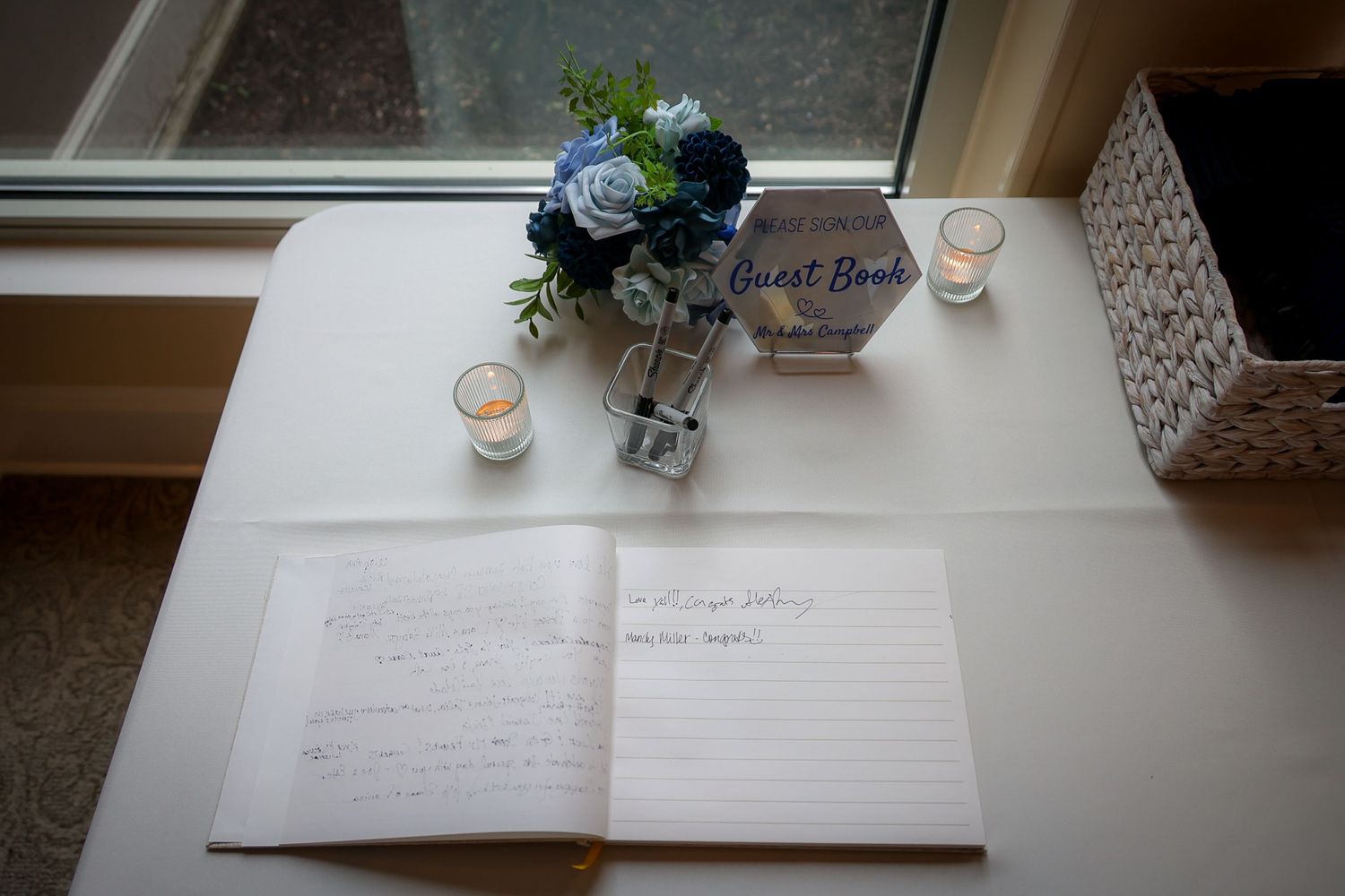 Open wedding guest book on white table with blue floral arrangement, candles, and decorative guest book box by window.