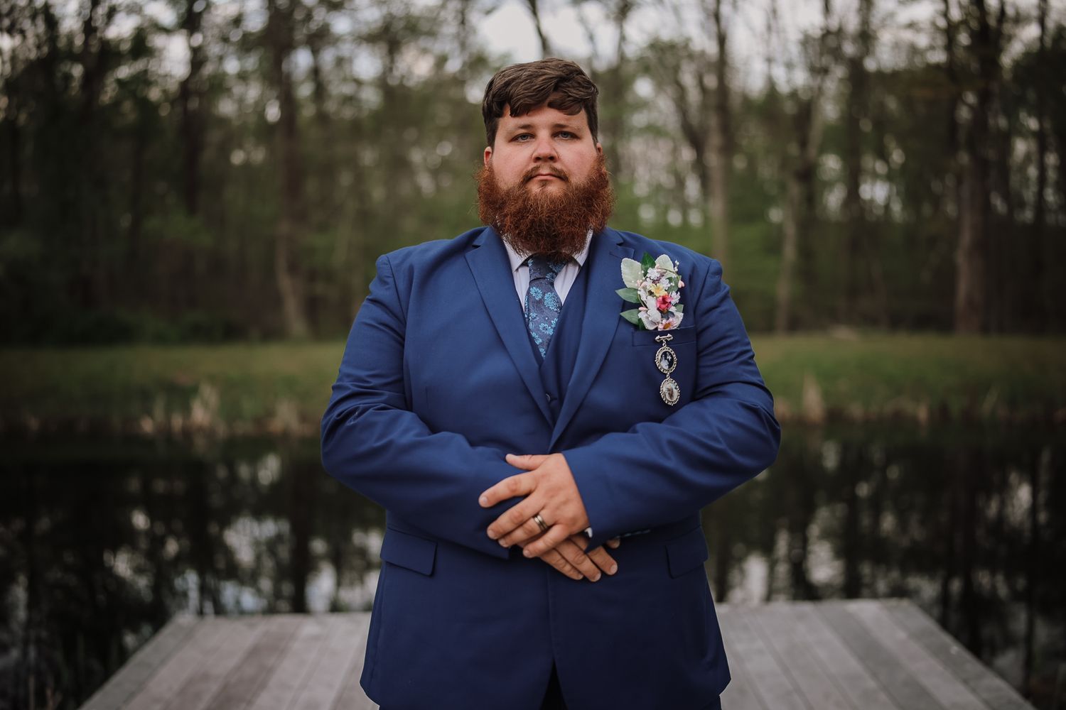Man in blue suit with floral boutonniere stands on wooden dock near lake, posing with arms crossed.