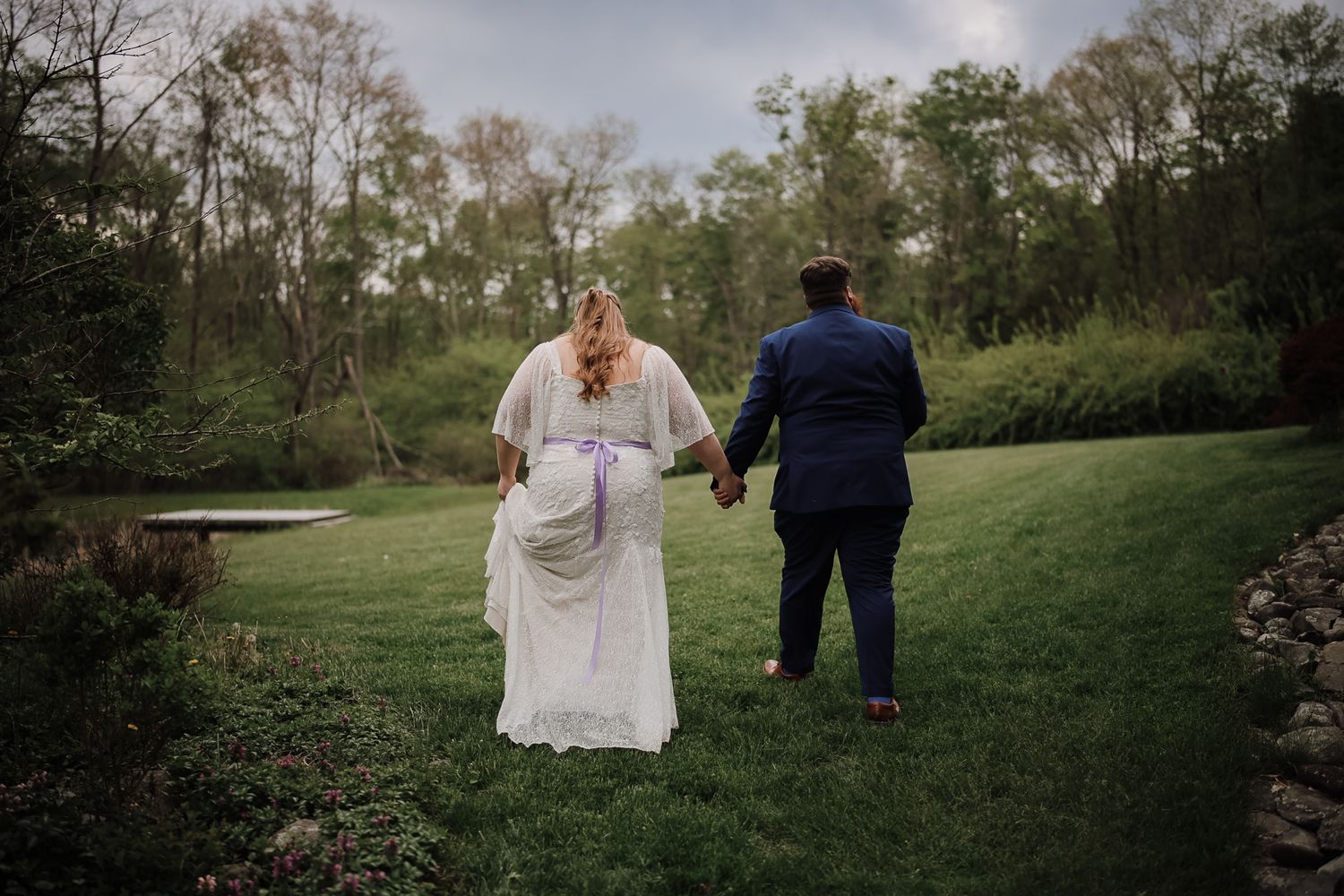 Bride and groom holding hands walking toward a lush green garden pond on their wedding day.