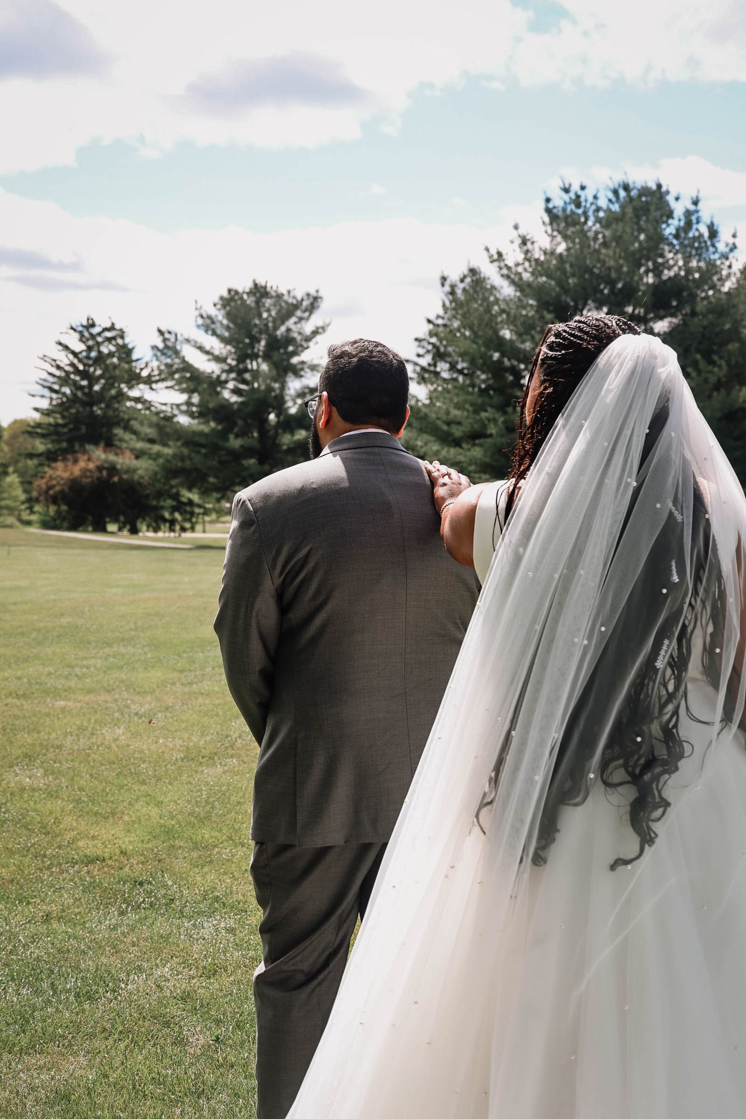 A bride and groom walk hand in hand across a lush green lawn, viewed from behind, with trees and cloudy sky ahead.