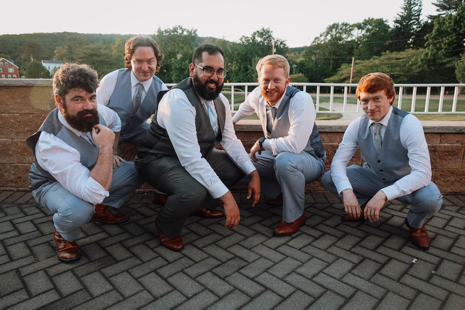 Five groomsmen in gray vests and light blue shirts pose playfully while crouching on a stone terrace at a wedding.