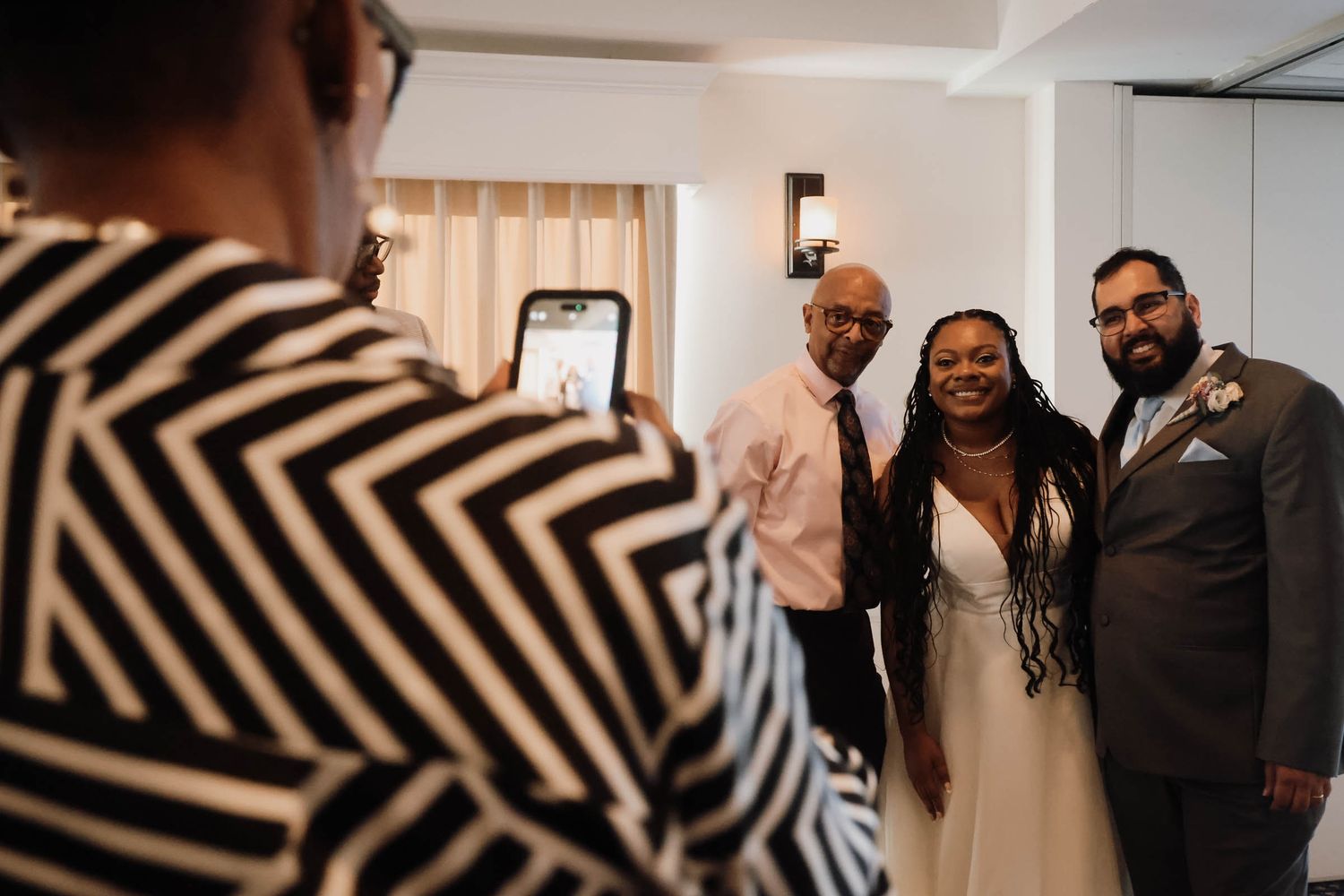 Wedding couple poses for photo with older gentleman as guest captures moment on smartphone in bright indoor venue.