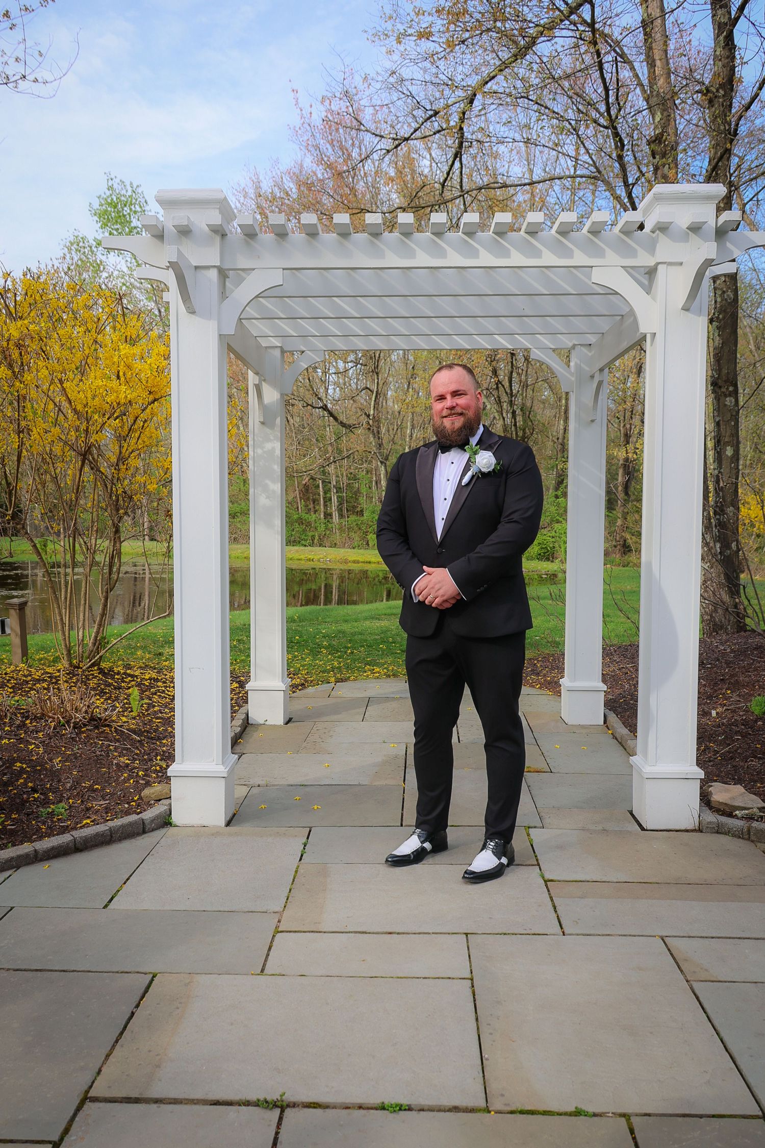 Groom in black tuxedo with white boutonniere stands under white pergola on stone patio surrounded by lush greenery.