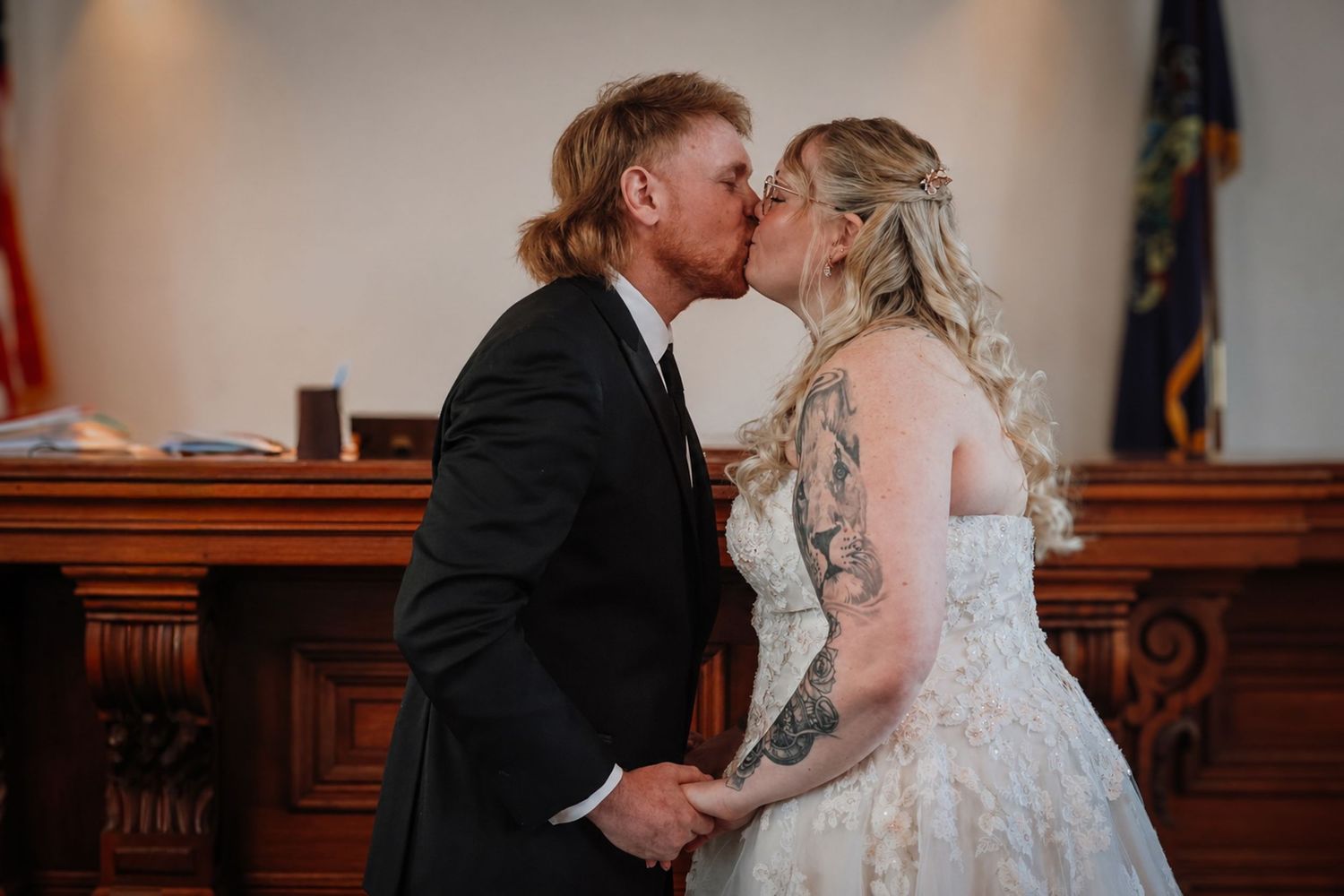 A bride and groom share their first kiss during an intimate courthouse wedding ceremony, bride wearing lace gown with tattoos visible.