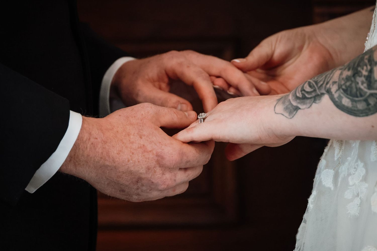 Groom placing a diamond ring on bride's tattooed hand during wedding ceremony ring exchange.