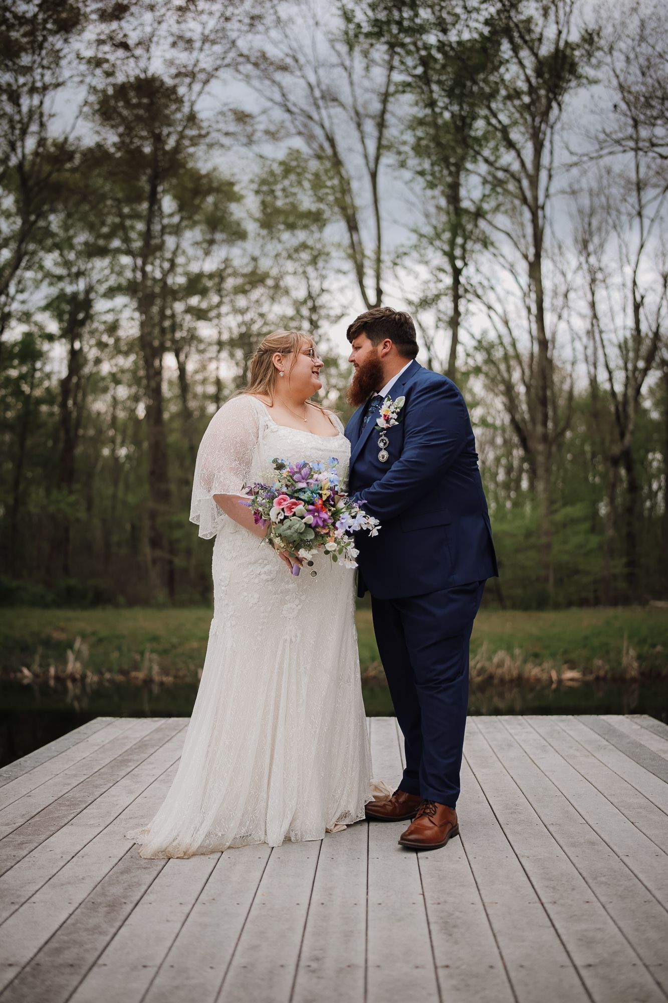 Bride and groom share a romantic moment on a wooden dock surrounded by trees, bride holding a colorful wildflower bouquet.