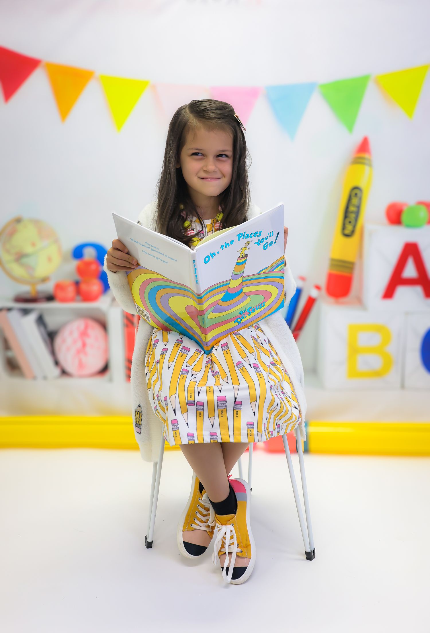 A young girl sits on a chair reading a Dr. Seuss book in a colorful classroom-themed backdrop with ABC decorations.