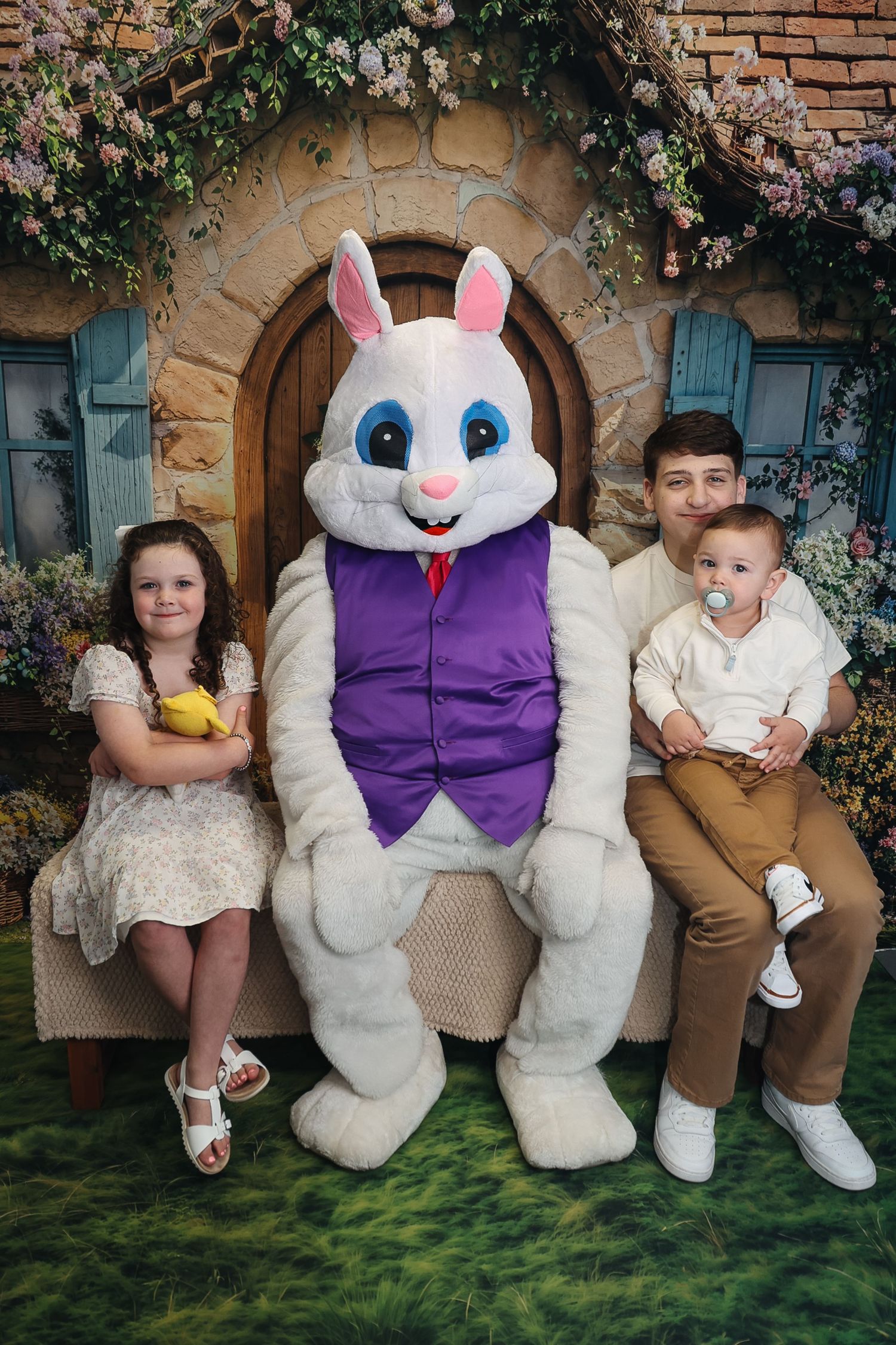 Three children posing with Easter Bunny mascot in front of decorated cottage backdrop for holiday photo.