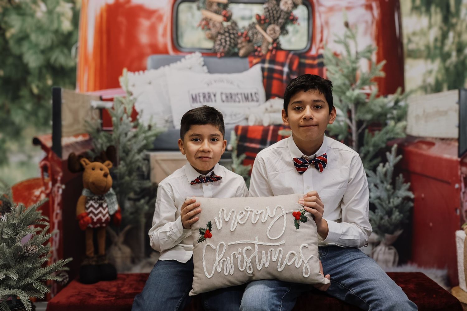 Two boys in white shirts and bow ties hold a Merry Christmas pillow in a festive holiday photo setting.