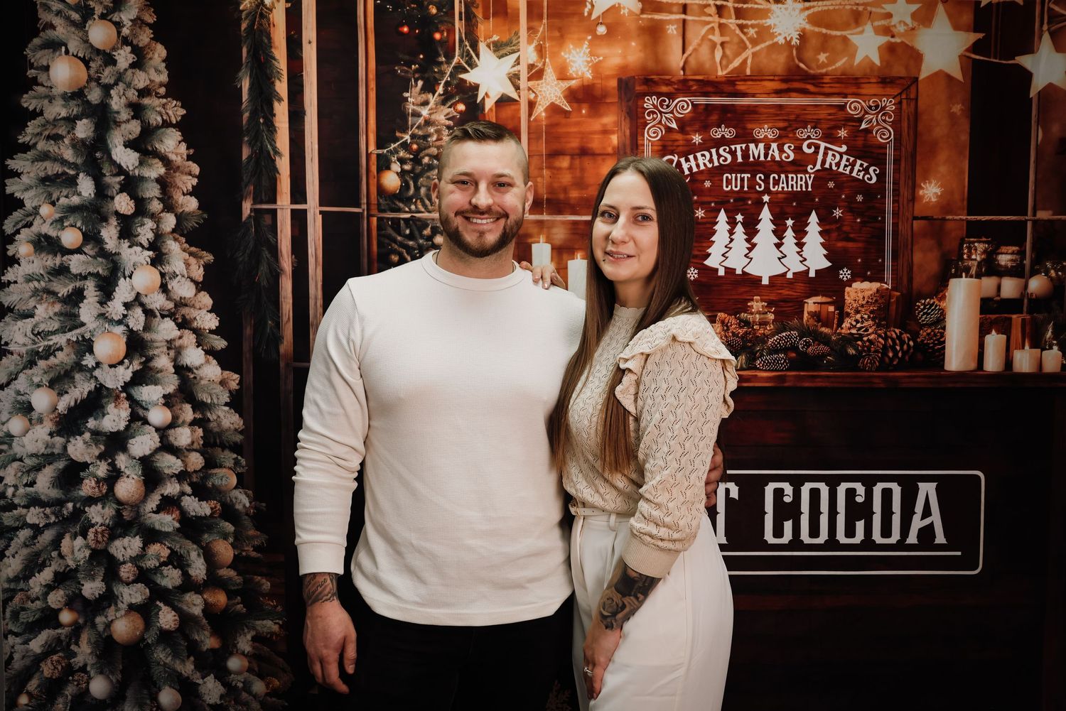 Couple posing together in front of a festive Christmas backdrop with holiday decorations and a hot cocoa sign.