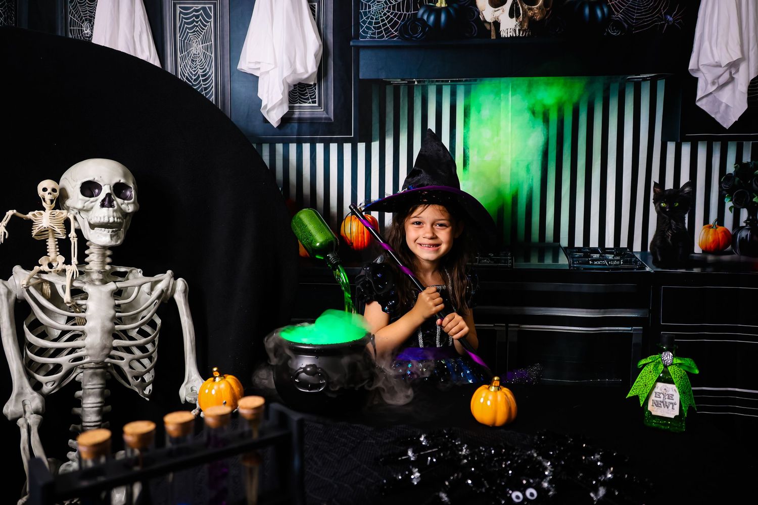 Young girl in witch costume with hat poses among Halloween decorations including skeleton, pumpkins, and cauldron.
