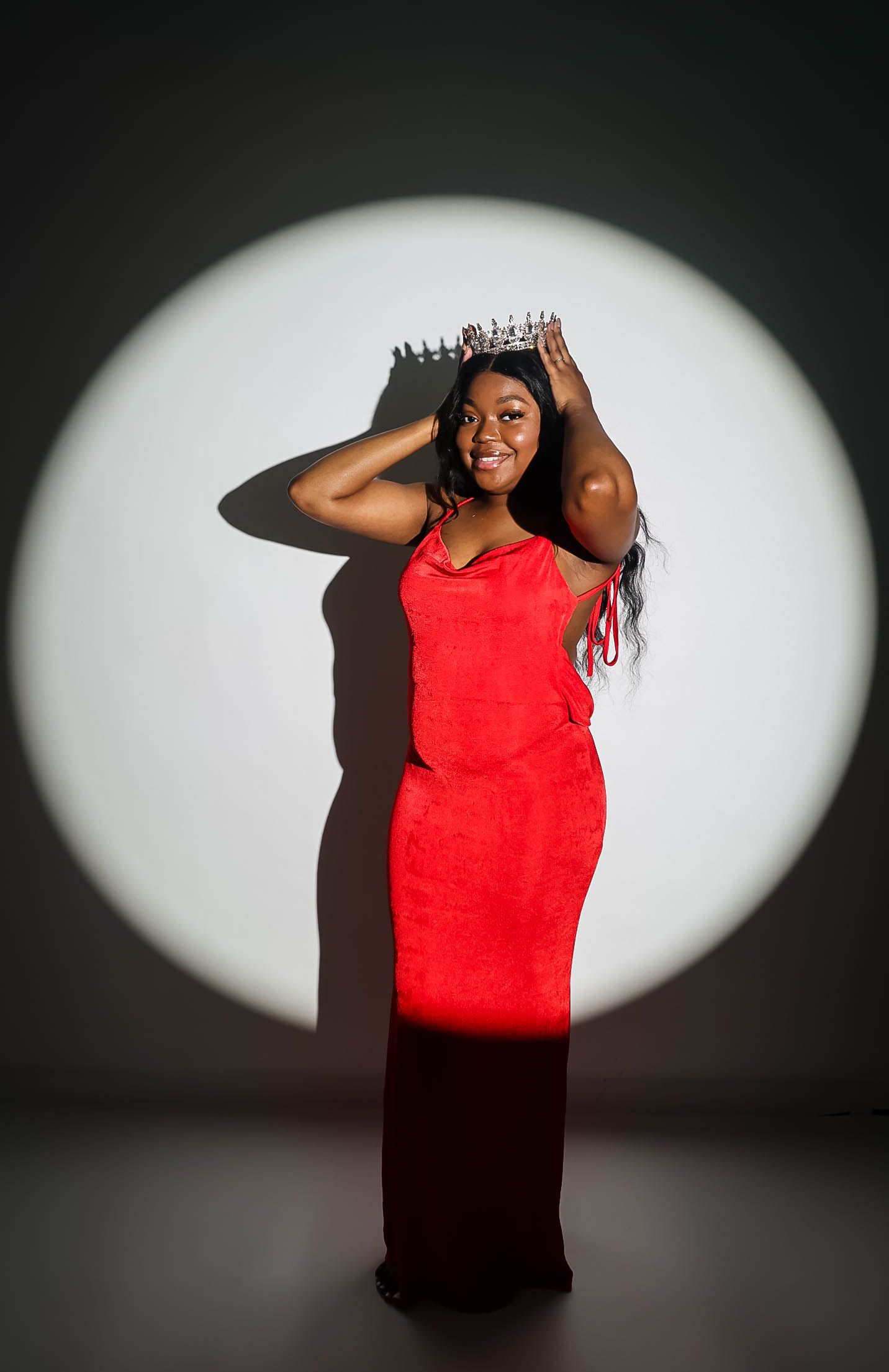 A smiling woman in a red gown holds a crown on her head, posed in front of a circular spotlight backdrop.