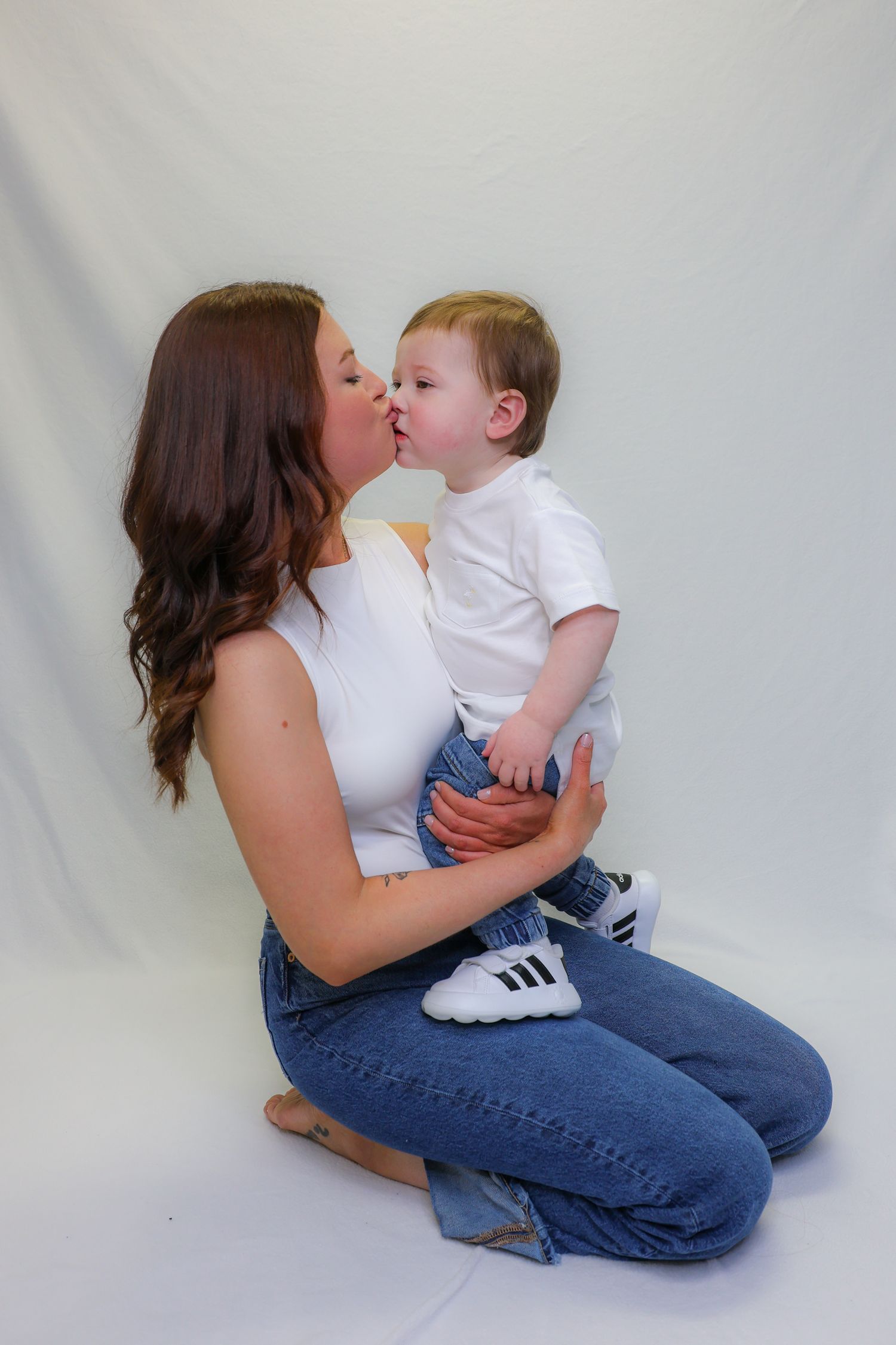 A young mother in jeans and white top kisses her baby boy in a white studio setting.