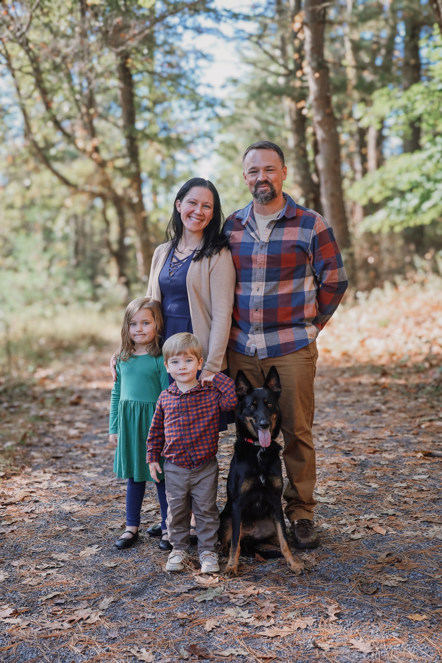 Family with two young children and a dog pose together on fallen autumn leaves in a wooded forest setting.