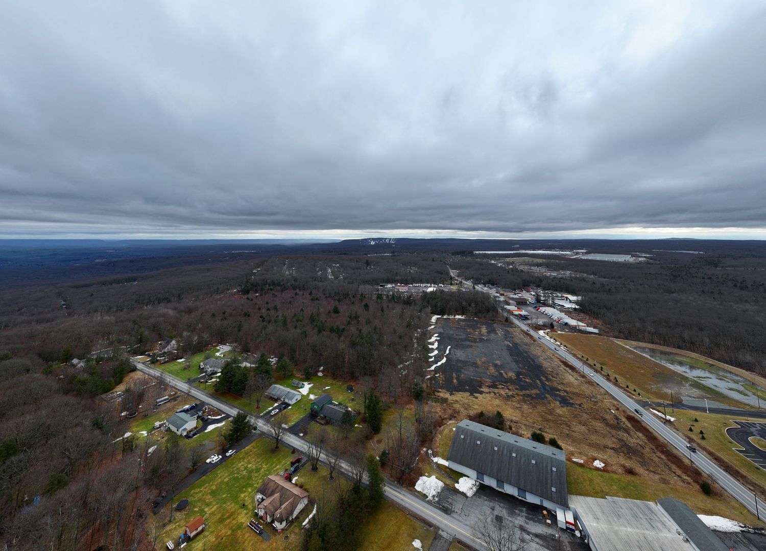 Aerial view of a small town surrounded by dense forests under overcast skies, with roads and buildings visible below.