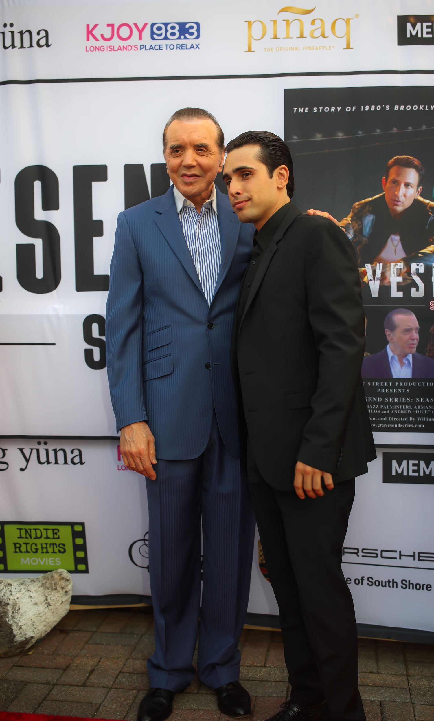 Two men in dark suits stand together at a promotional event with posters and signage in the background.