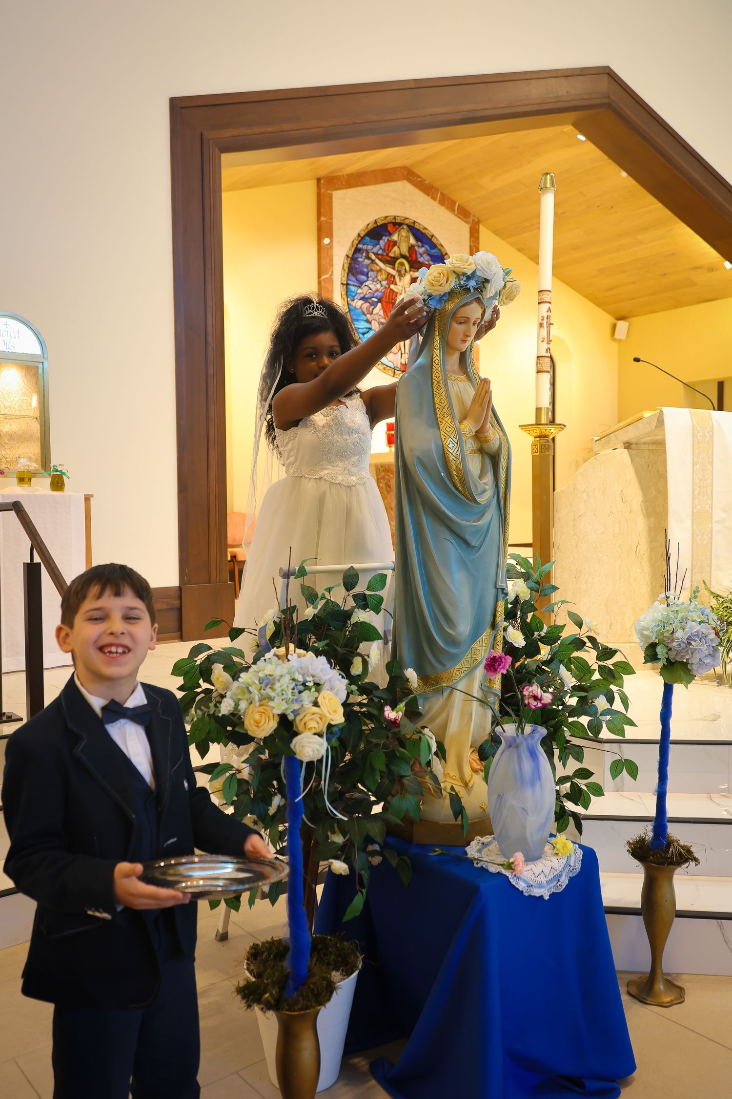 A religious ceremony in a church with a statue of Mary being crowned while a young person in formal attire stands nearby.