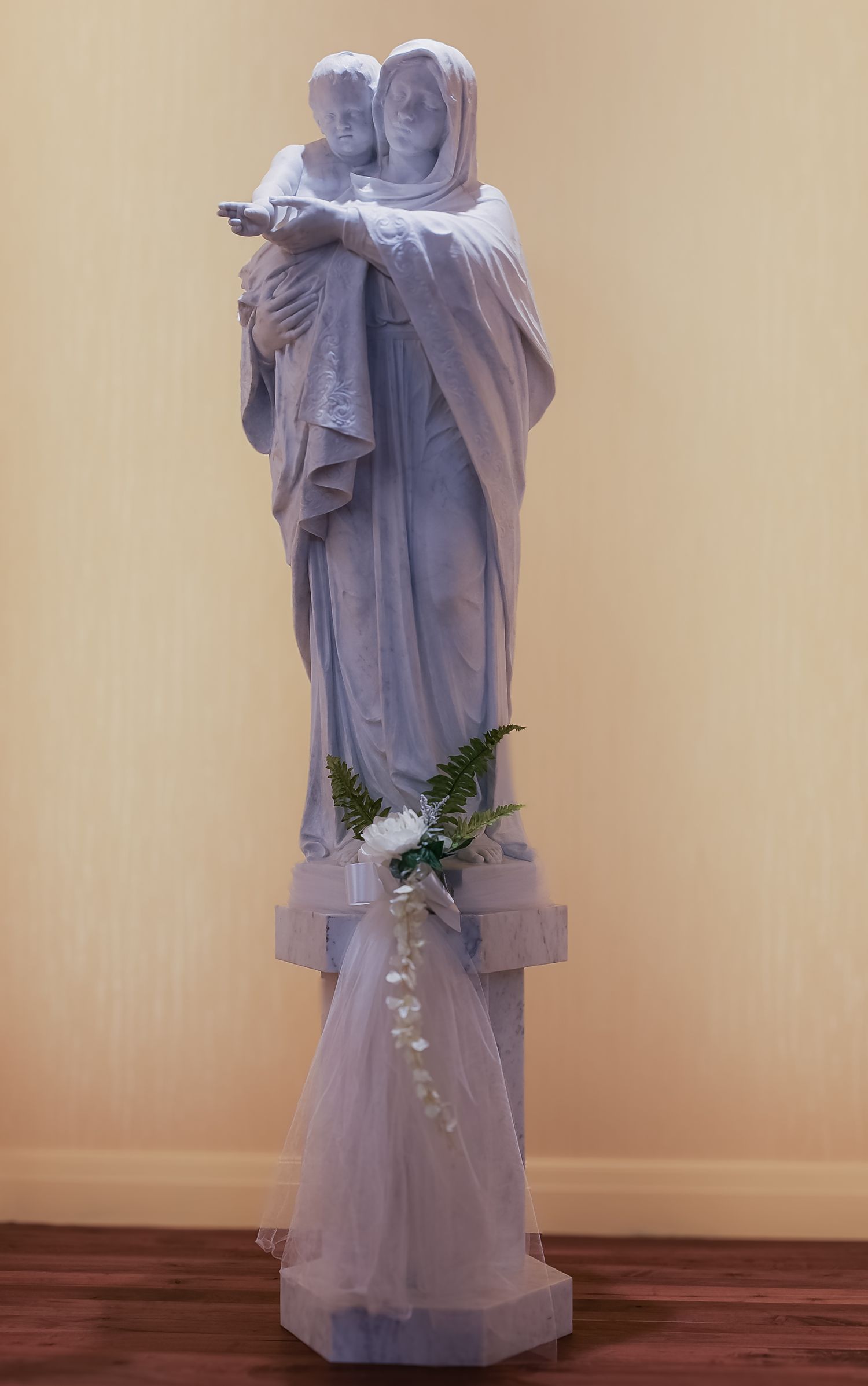 White religious statue adorned with delicate tulle and floral decorations stands on wooden floor against beige wall.