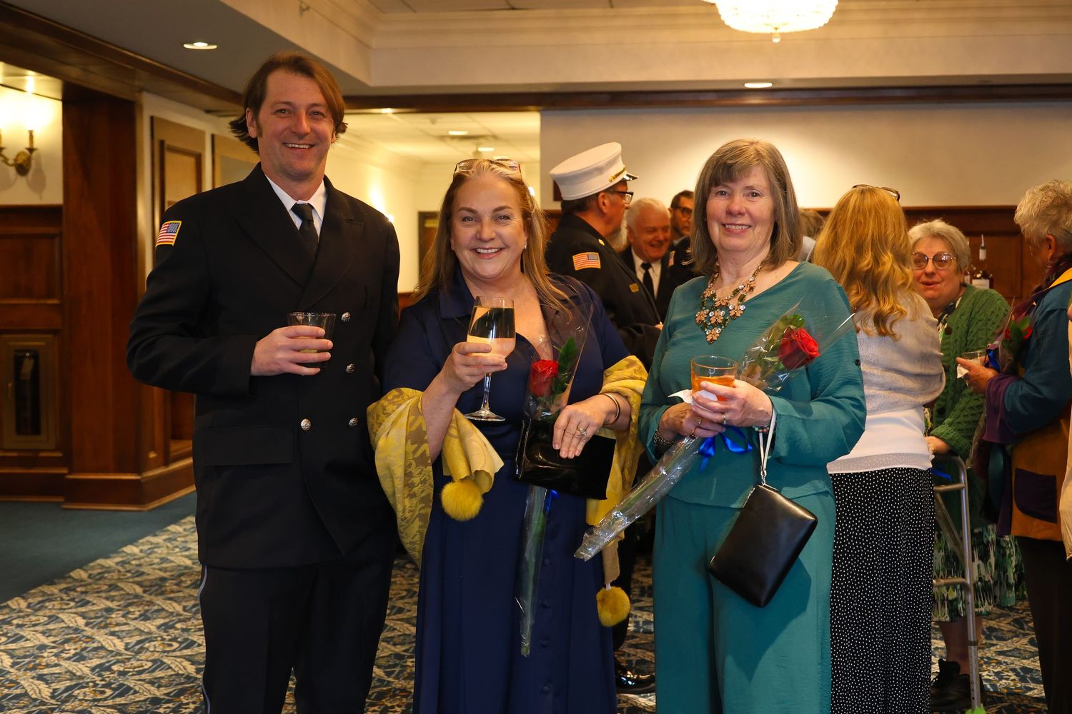A group of people in formal attire gather at an indoor event, holding drinks and socializing.