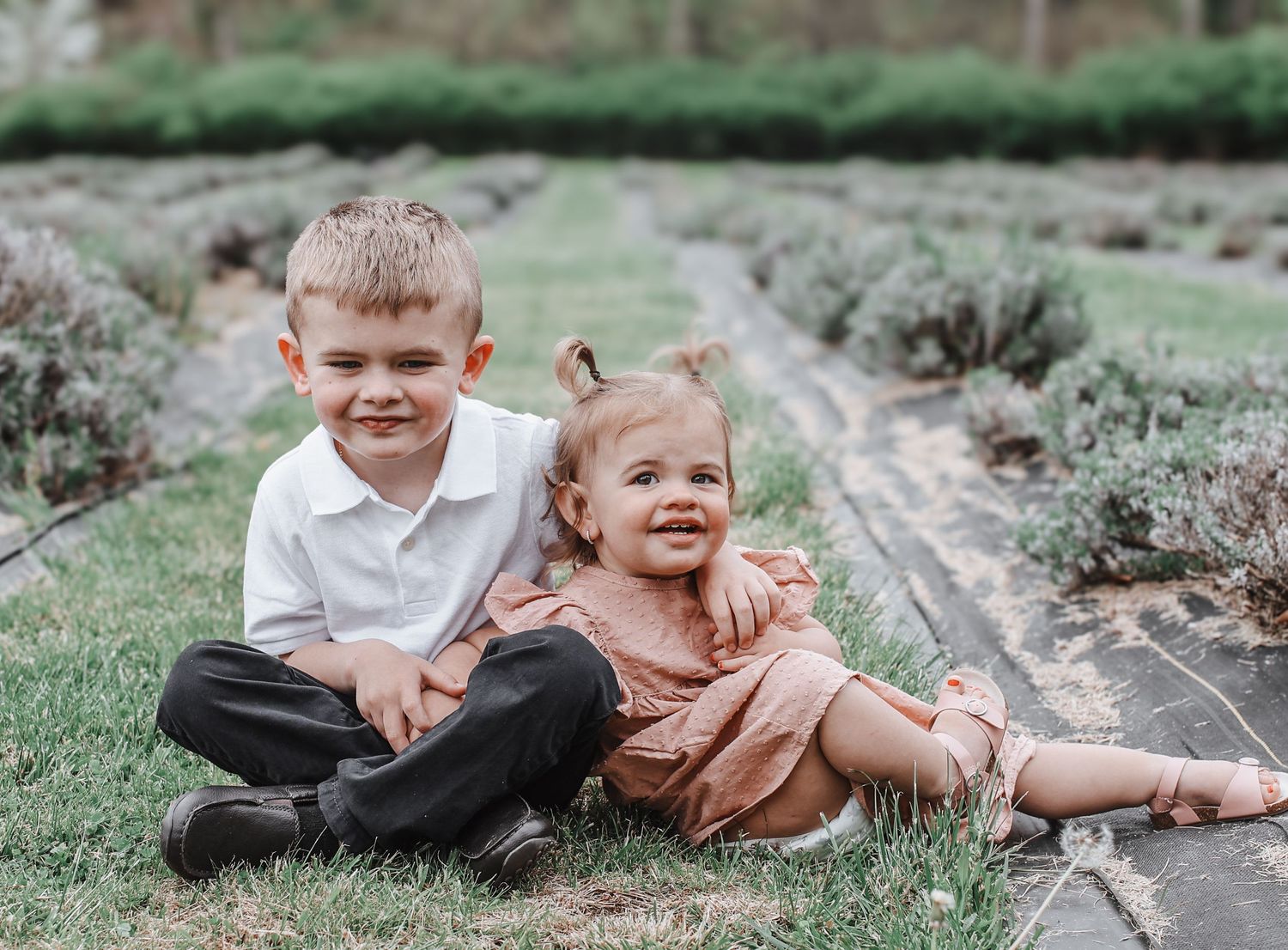 Two young children sit together outdoors on a garden path surrounded by green foliage.