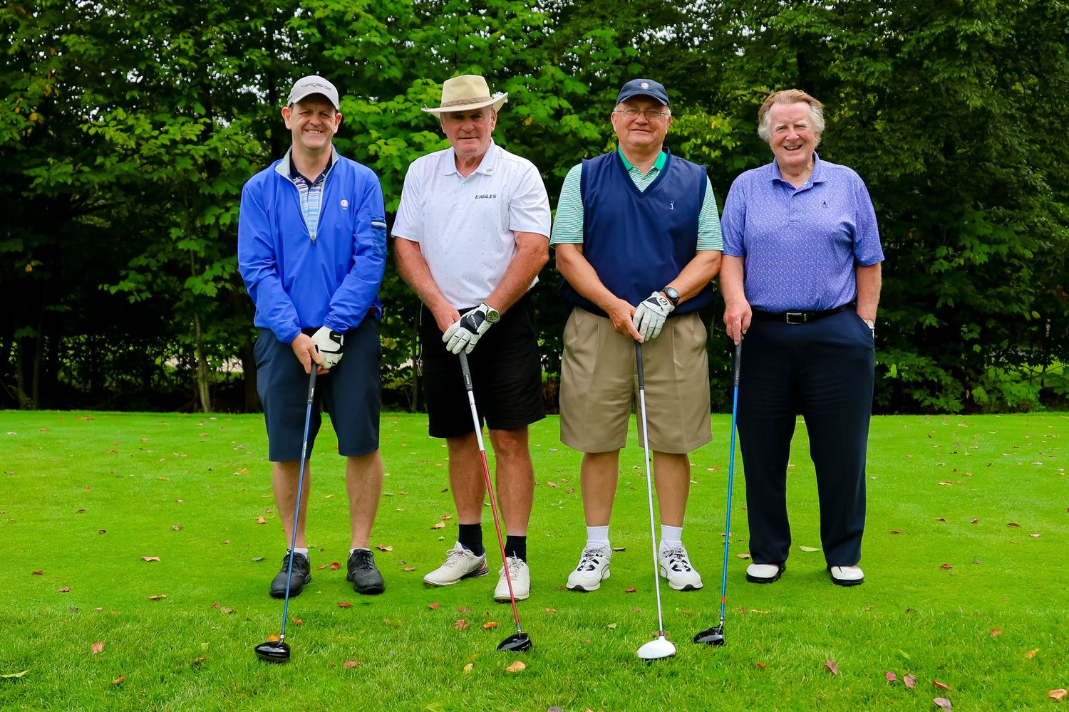 Four golfers stand together with their clubs on a lush green golf course surrounded by trees.
