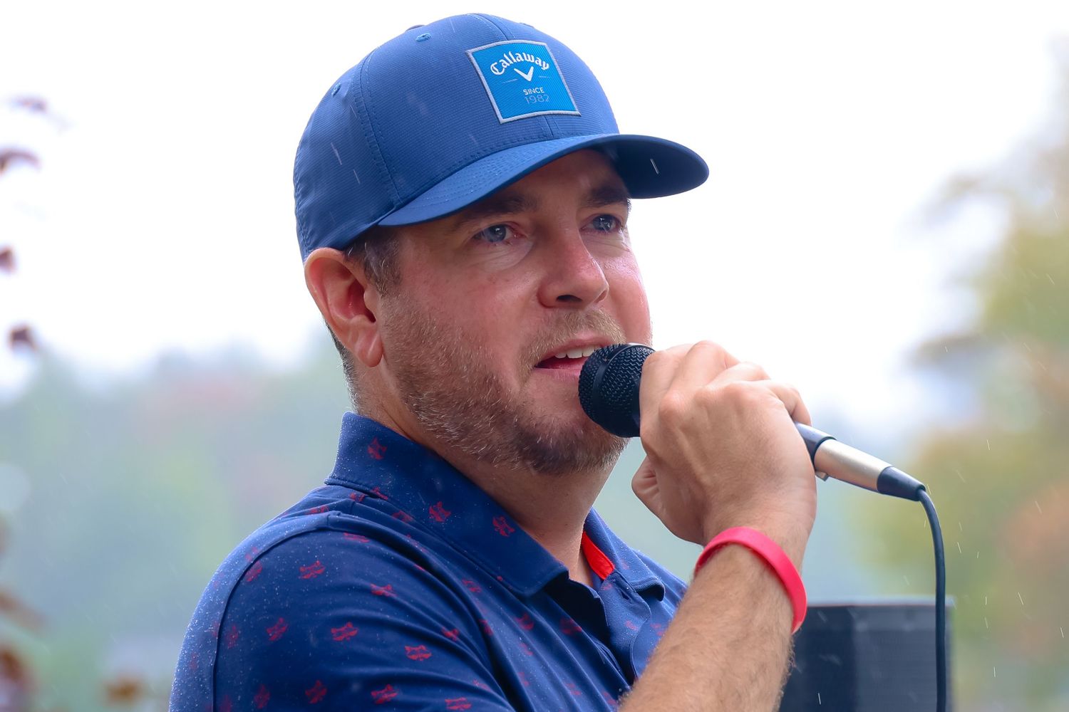 A performer wearing a blue baseball cap speaks into a microphone during an outdoor event.