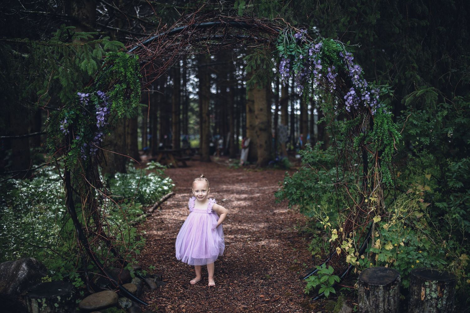 A child in a pink dress walks along a wooded forest path surrounded by tall trees and dappled sunlight.