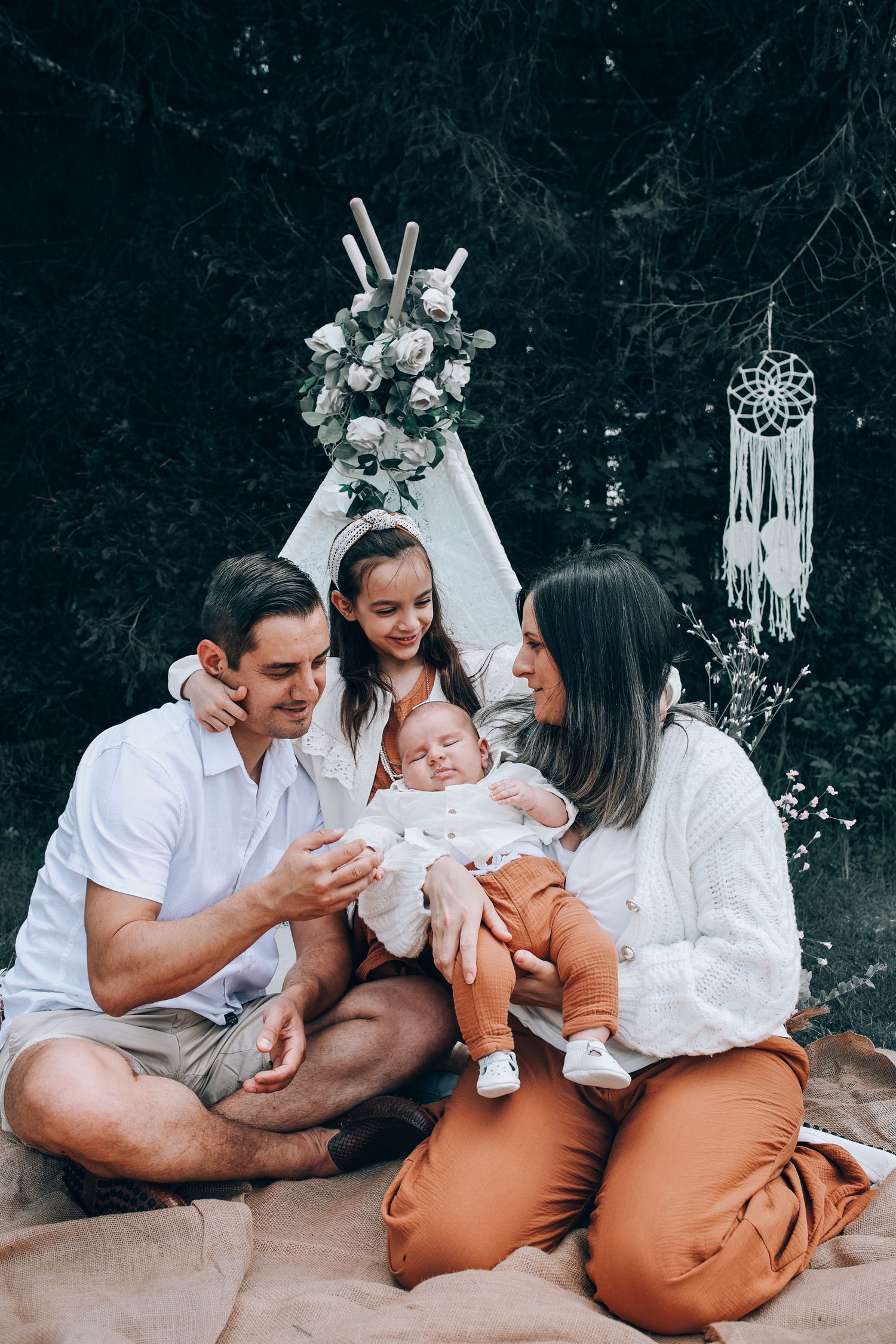 Family wearing white and brown outfits sits together on ground with boho dreamcatcher decor in outdoor setting.