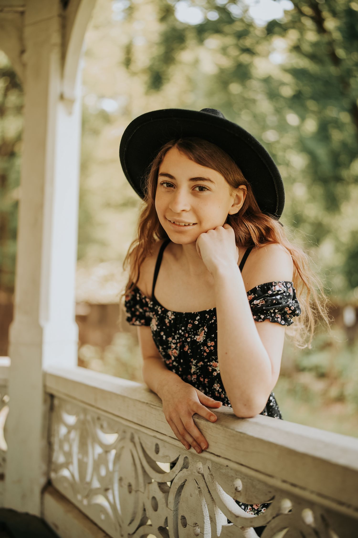 A child in a floral dress and black hat leans playfully on a white porch railing in soft natural lighting.