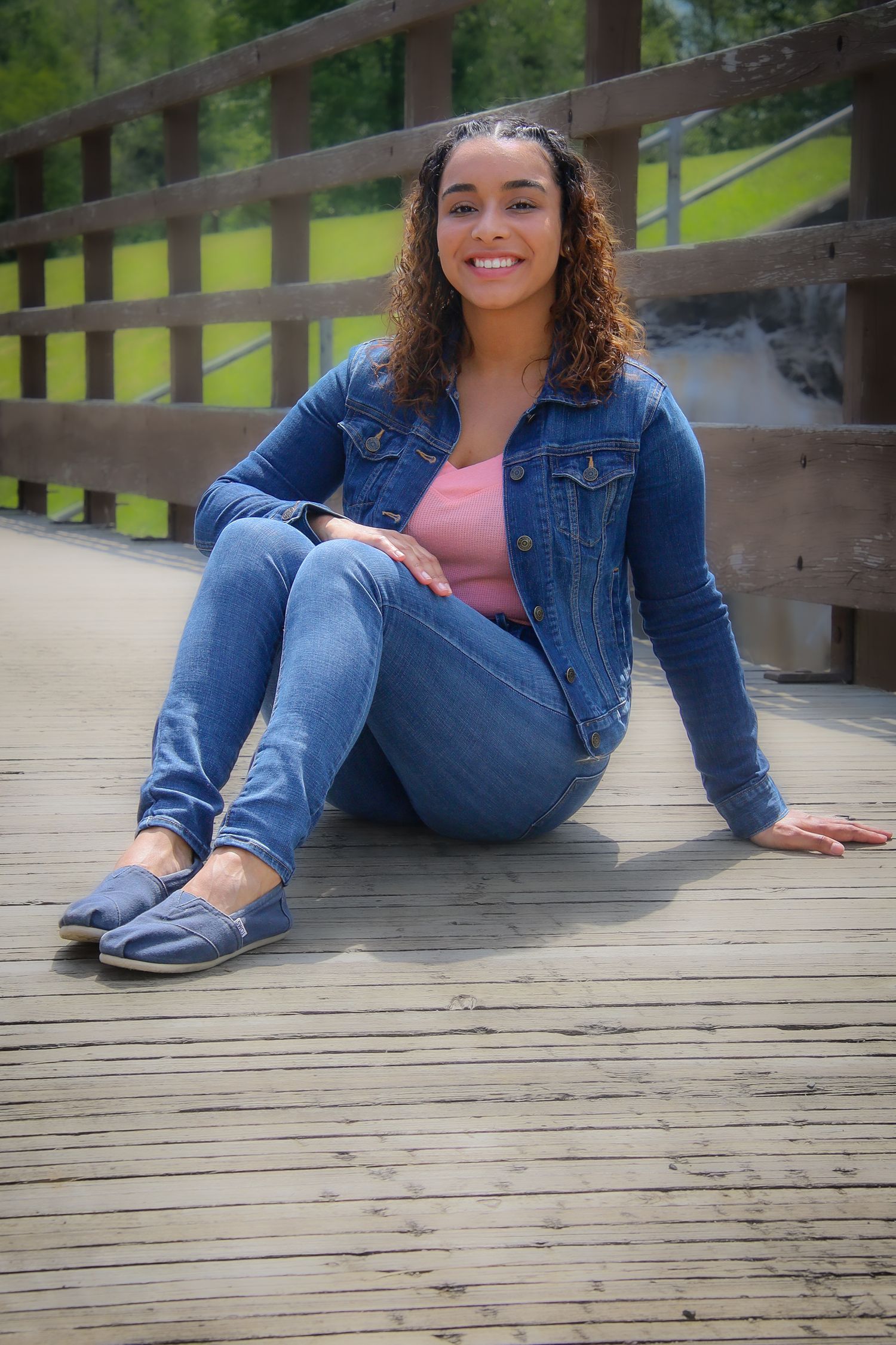 Someone in casual denim attire sits relaxed on a wooden bridge walkway surrounded by green scenery.