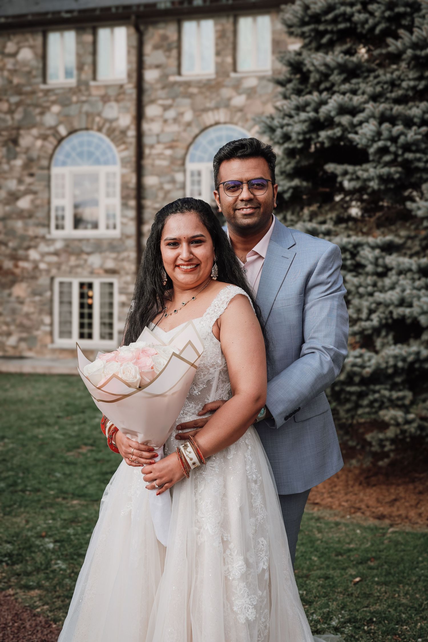 A couple in wedding attire poses together in front of a stone building with a bouquet of flowers.