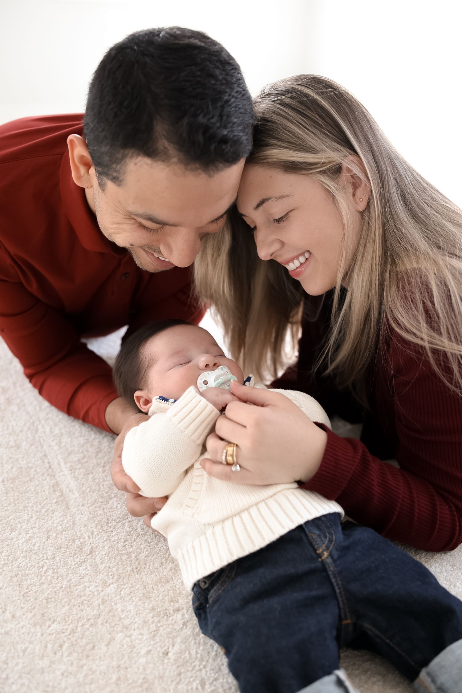 A couple in burgundy and cream outfits share intimate moments while laying on a white carpet.