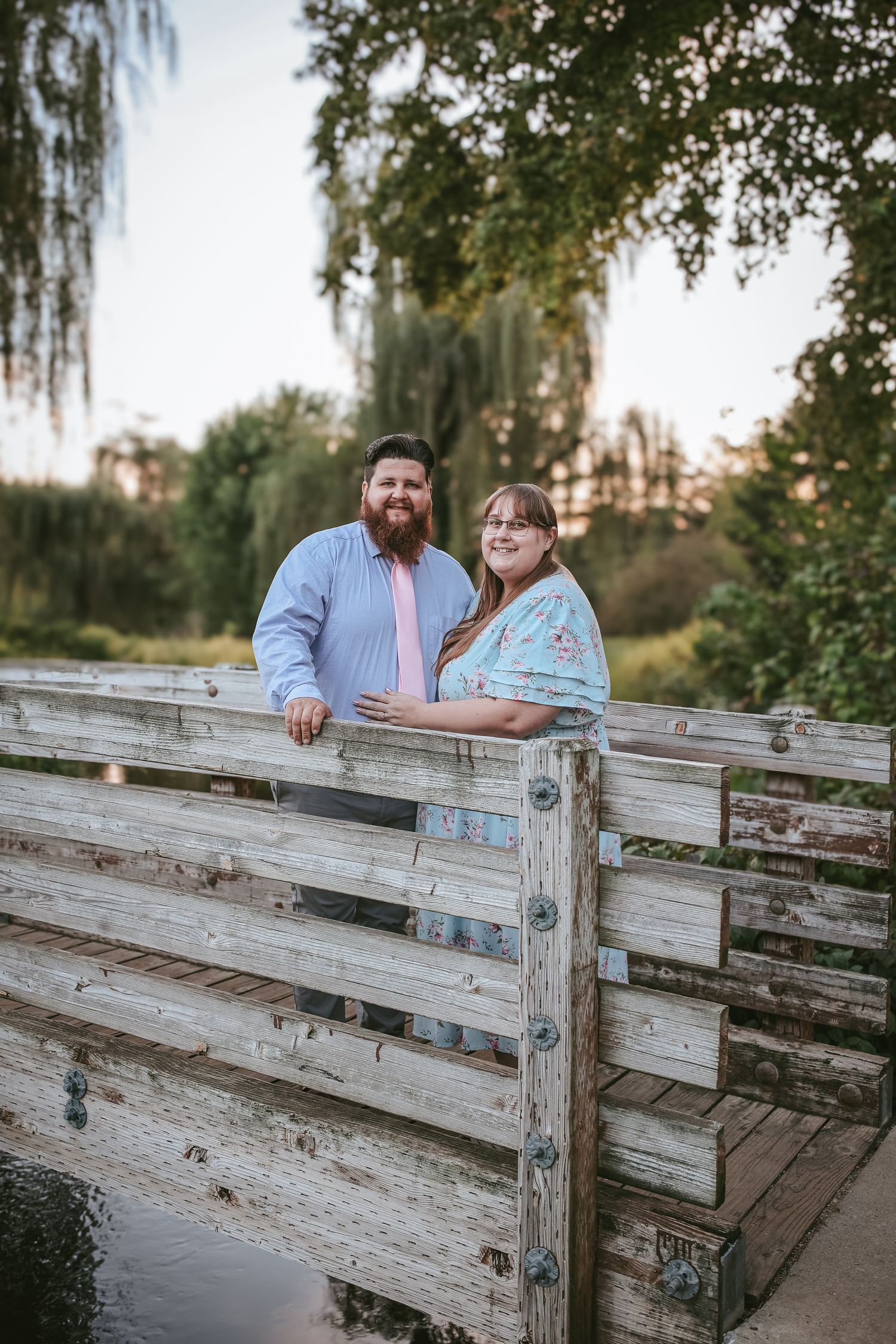 Couple sharing intimate moments on a wooden bridge surrounded by willow trees at sunset.