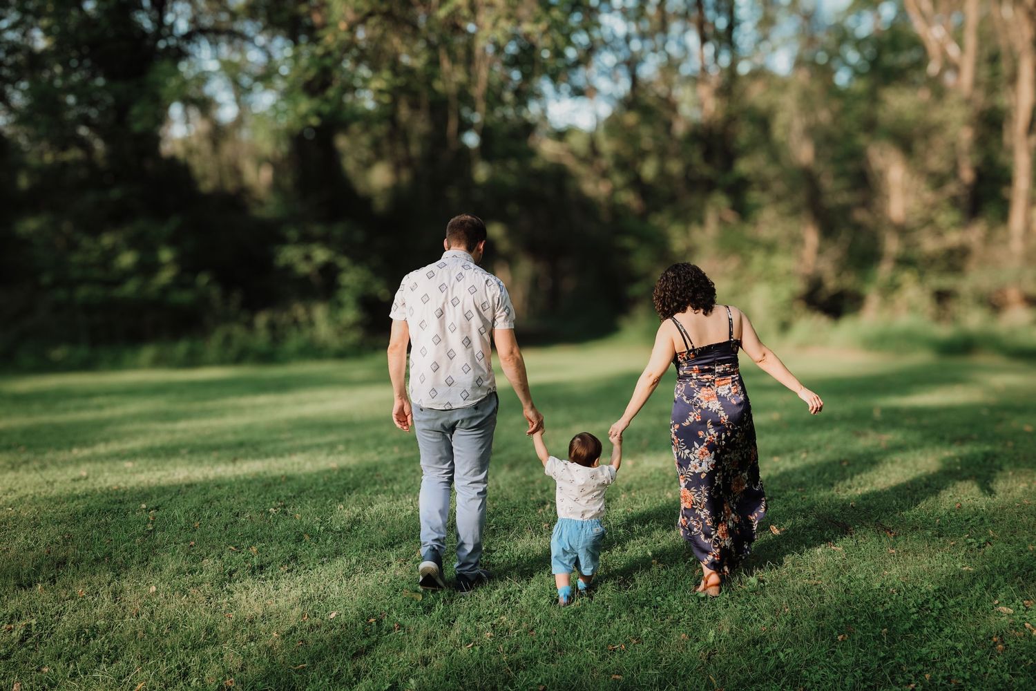 Family holding hands while walking together on a grassy lawn surrounded by trees on a sunny afternoon.