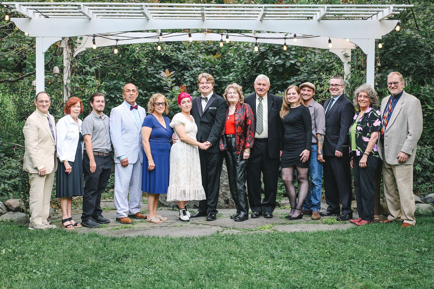 Large group of wedding guests pose together outdoors on grassy lawn during summer celebration.
