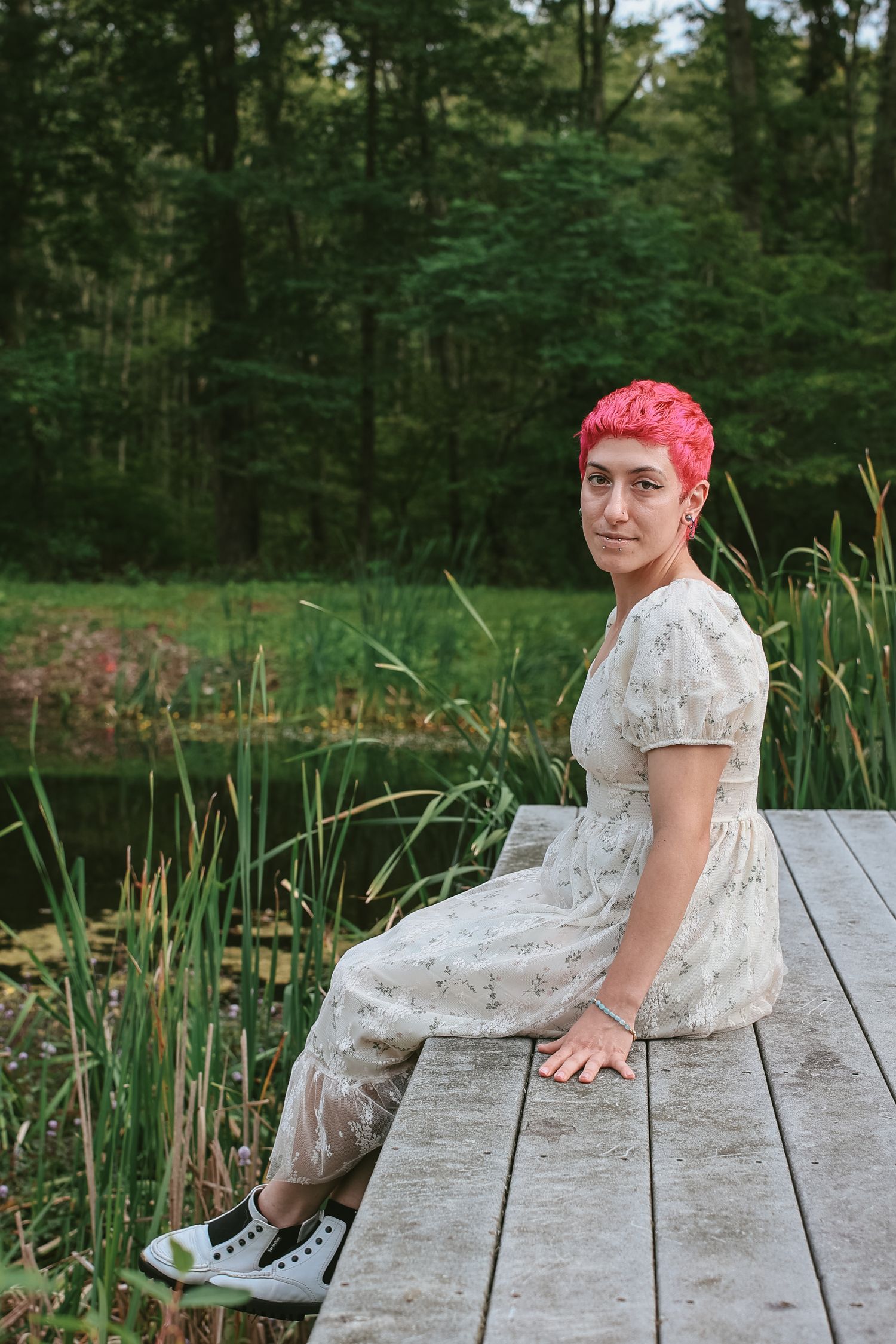 A person with bright pink hair wearing a white dress reclines on a wooden dock by a pond surrounded by greenery.