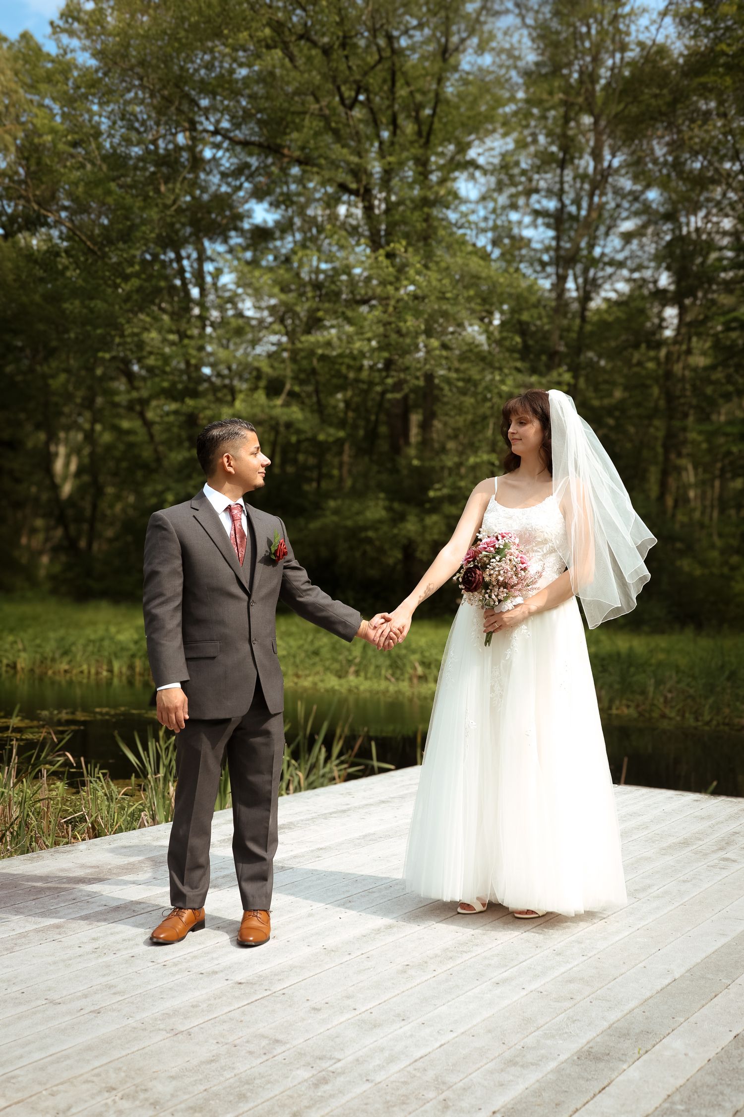A couple in wedding attire stands together on a wooden deck surrounded by lush green trees in a natural outdoor setting.