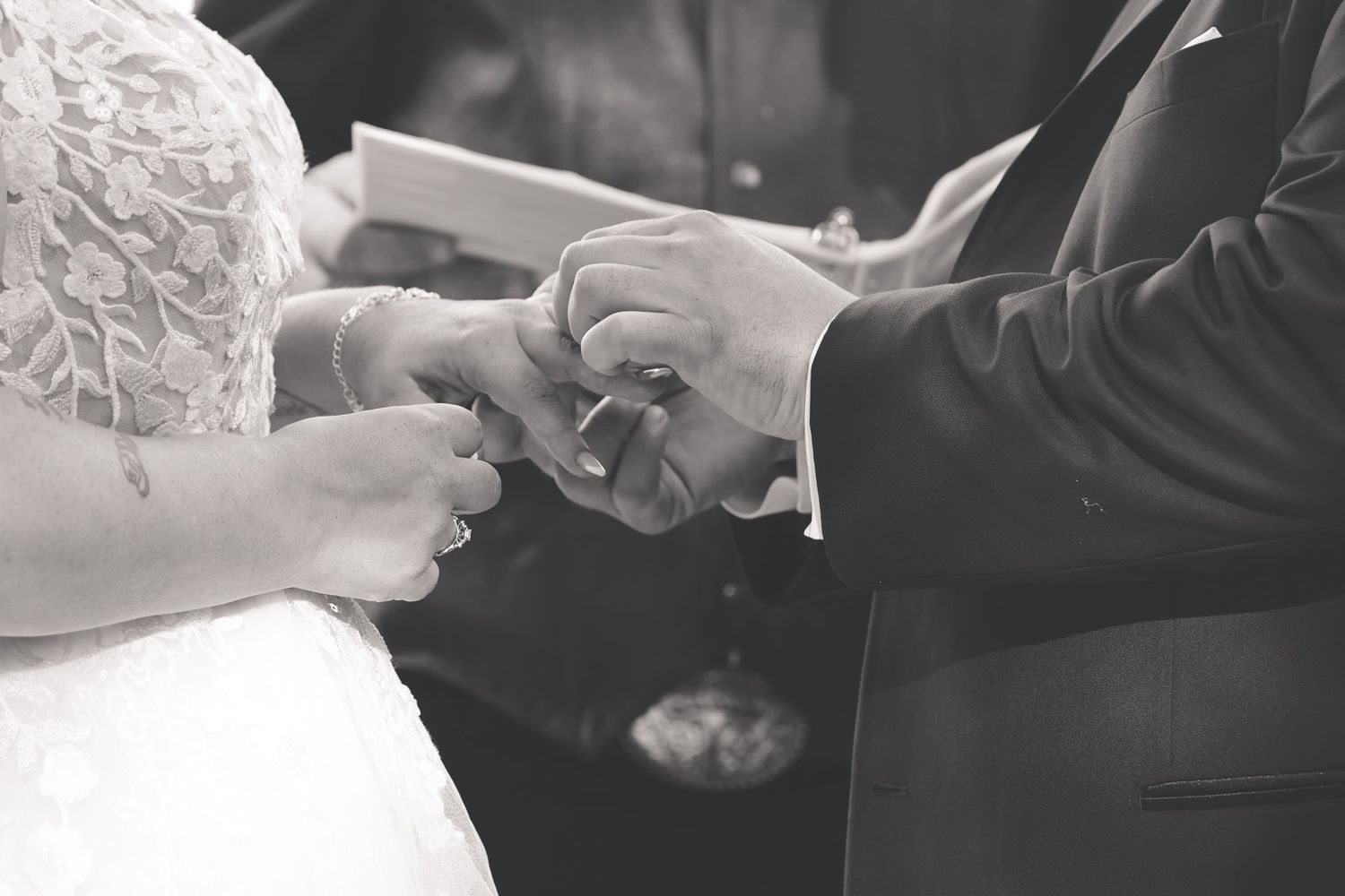 A close-up black and white photograph of hands exchanging wedding rings during a marriage ceremony.